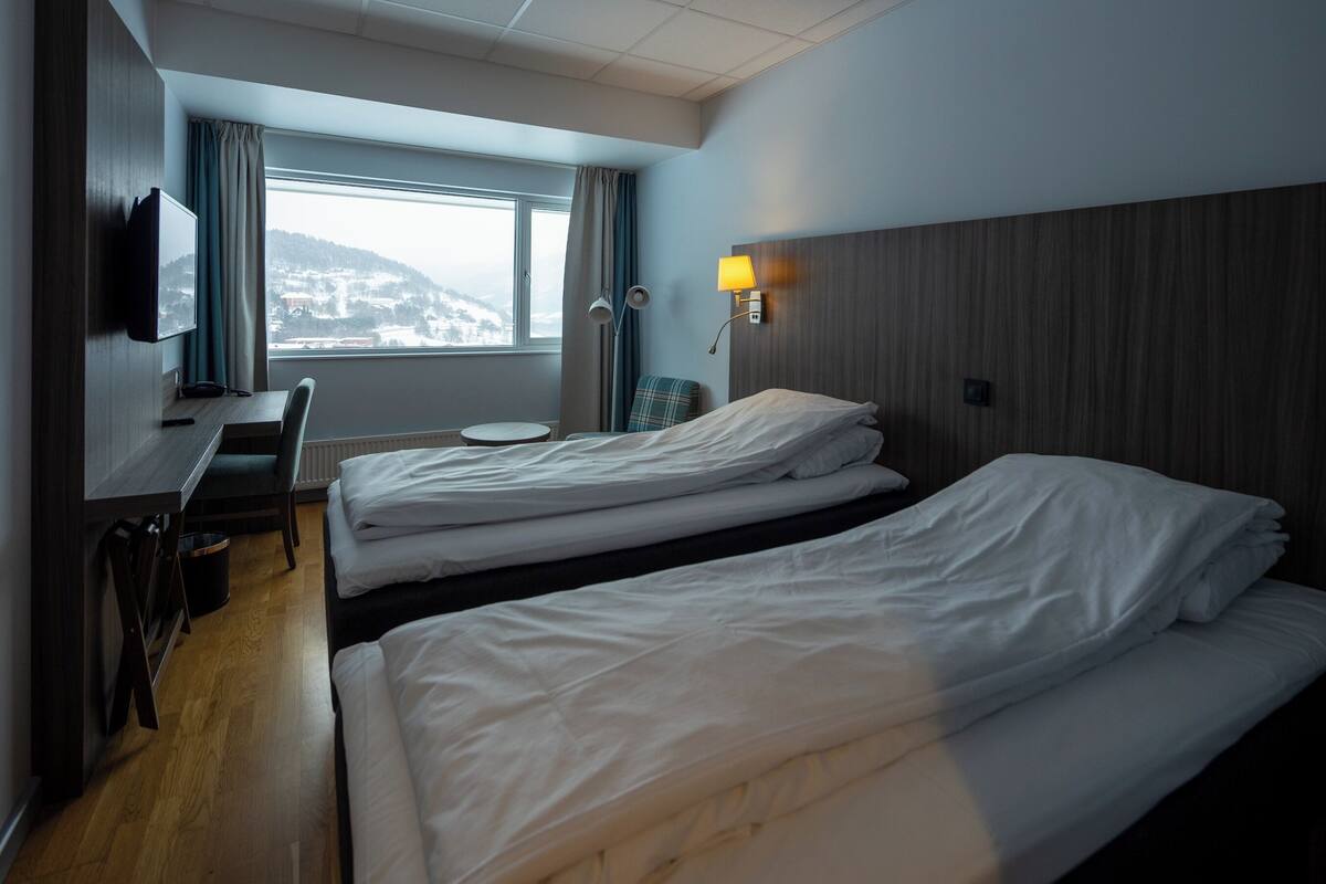 Ein Hotelzimmer mit zwei Betten, einem Schreibtisch, einem Sessel, einem Fernseher und einem Fenster mit Blick auf eine verschneite Landschaft.