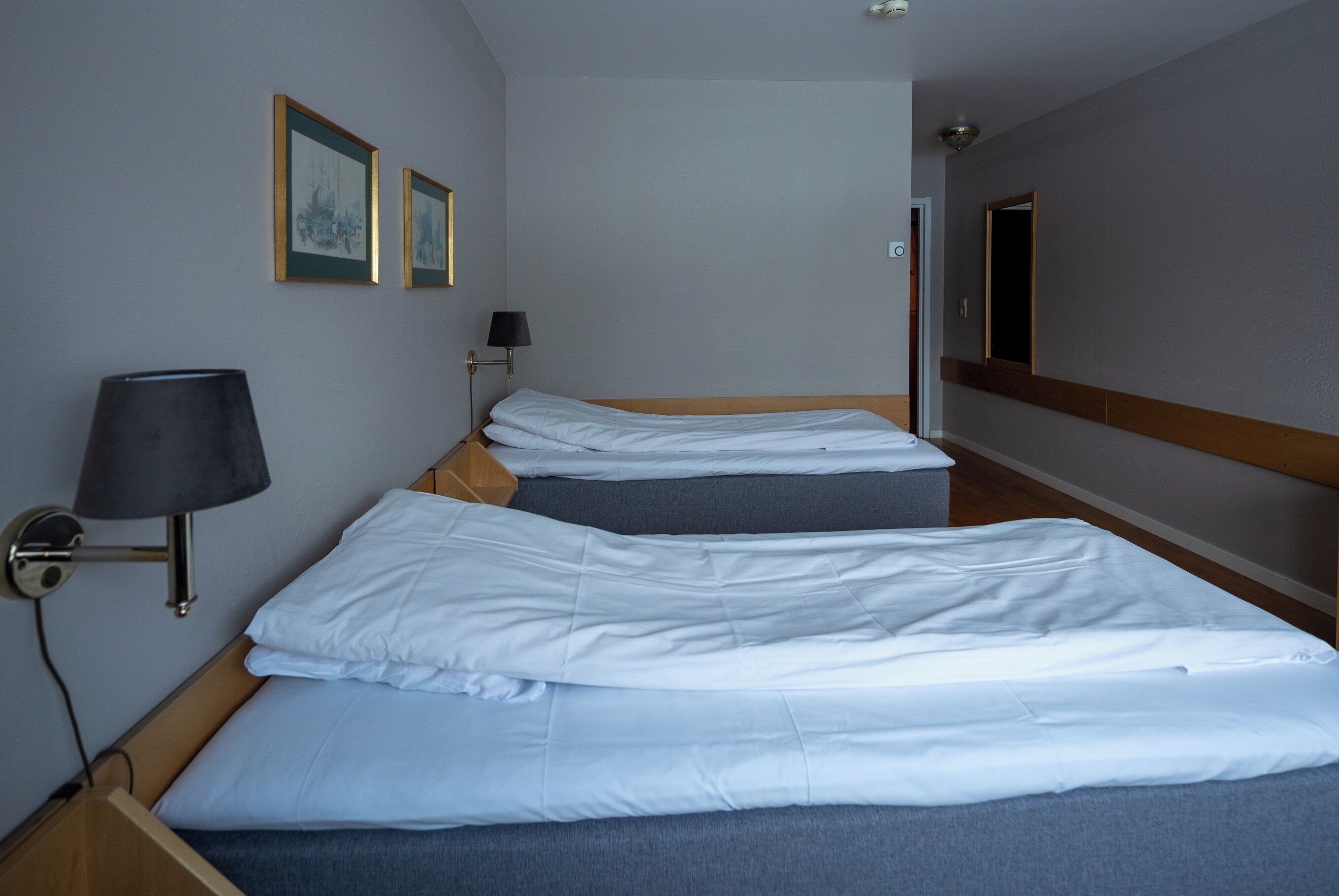 Habitación de hotel con dos camas individuales, una mesita de noche, una lámpara y cuadros en la pared.
