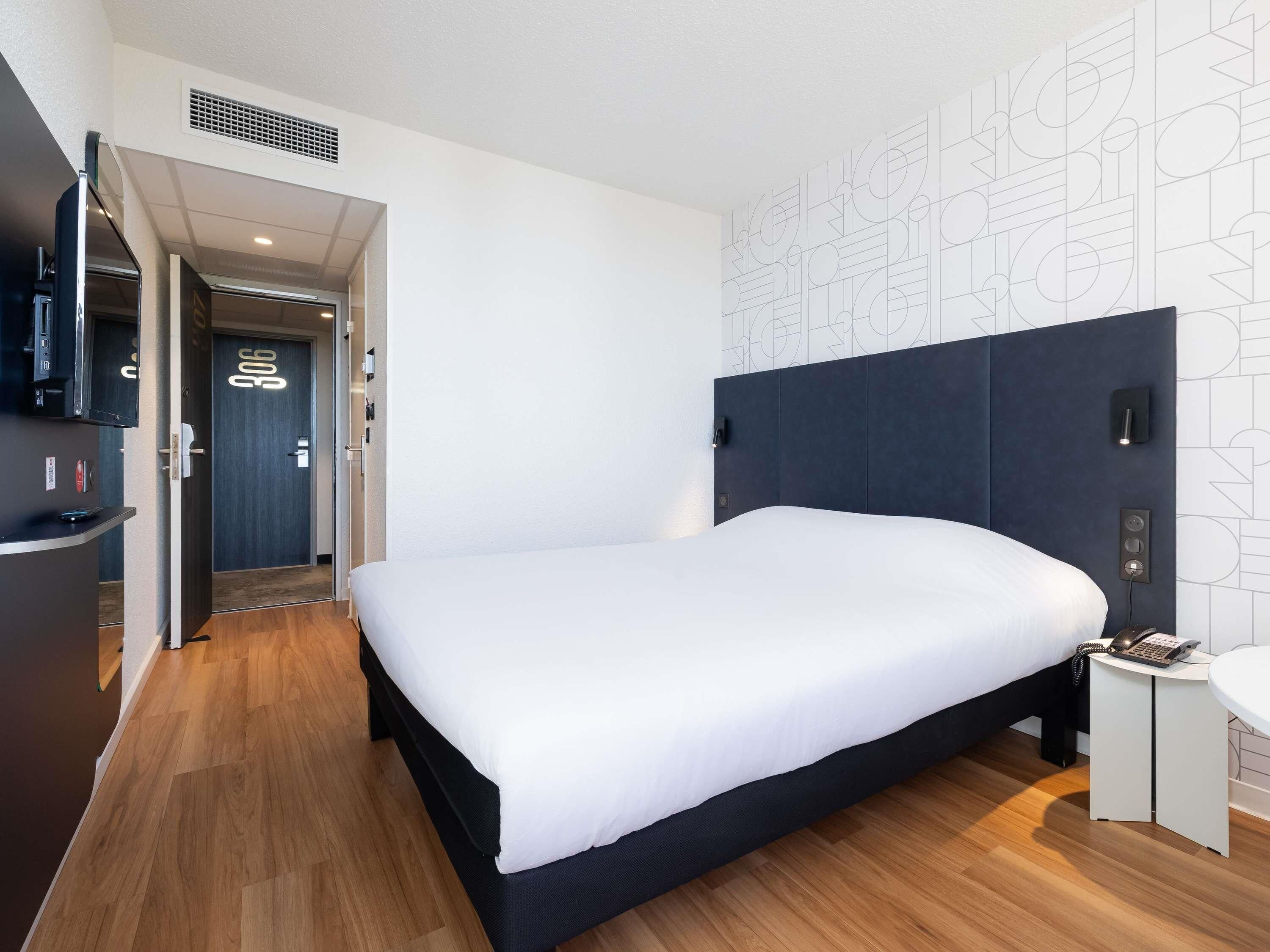Habitación de hotel moderna con una cama grande, una mesita de noche, un escritorio con teléfono y una puerta que da a otra habitación.