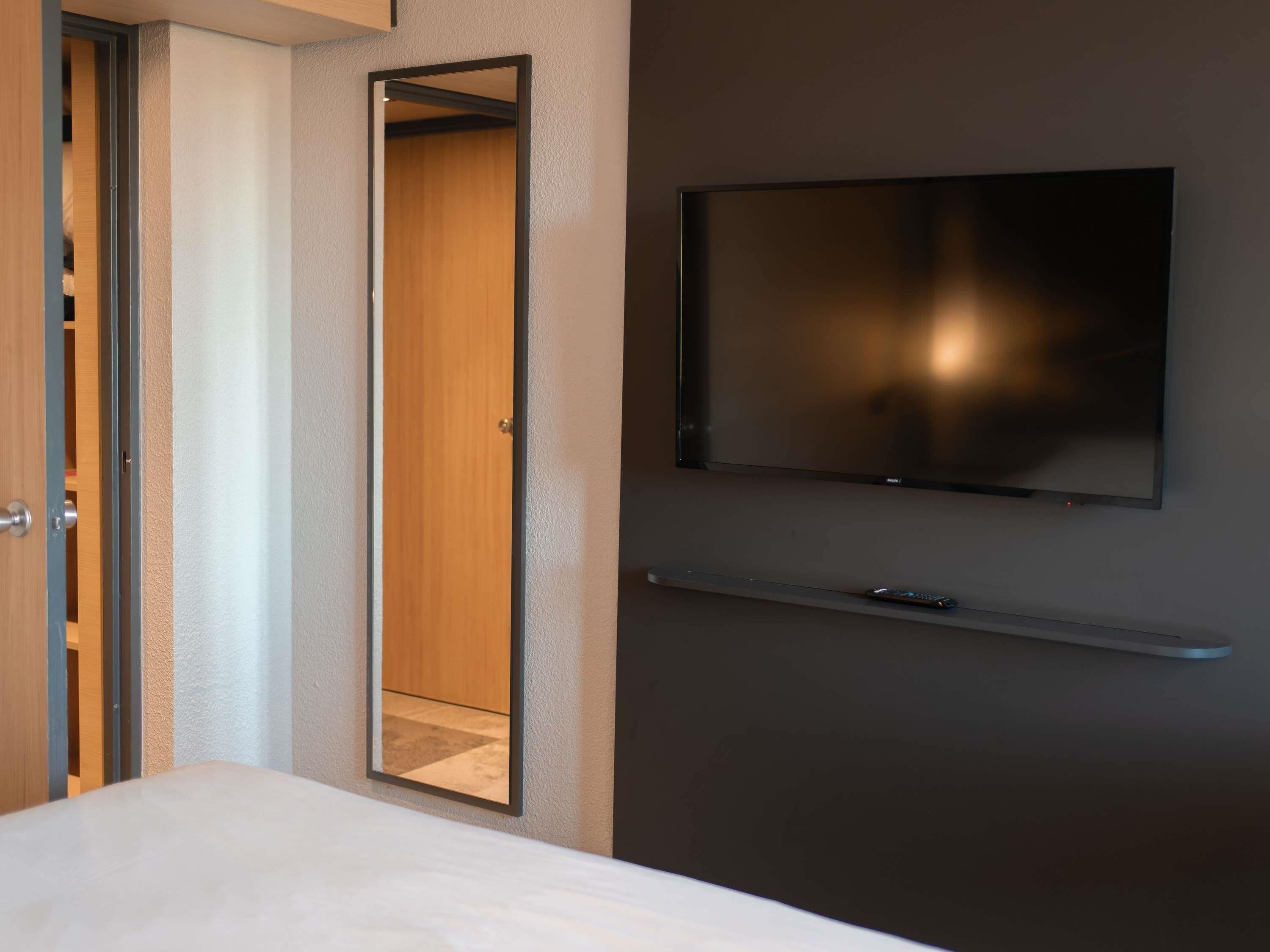 Ett hotellrum med en säng, en garderob, en spegel, en tv som är monterad på väggen och en fjärrkontroll som ligger på sängen.