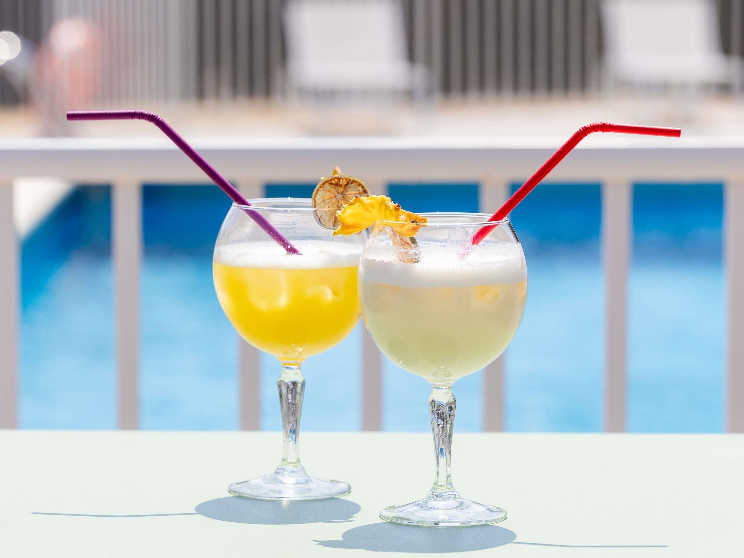 Two cocktails with straws and garnishes on a table by a pool.