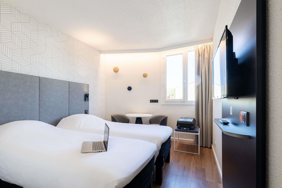 A hotel room with two beds, a desk with a laptop, a TV, and a window with curtains.