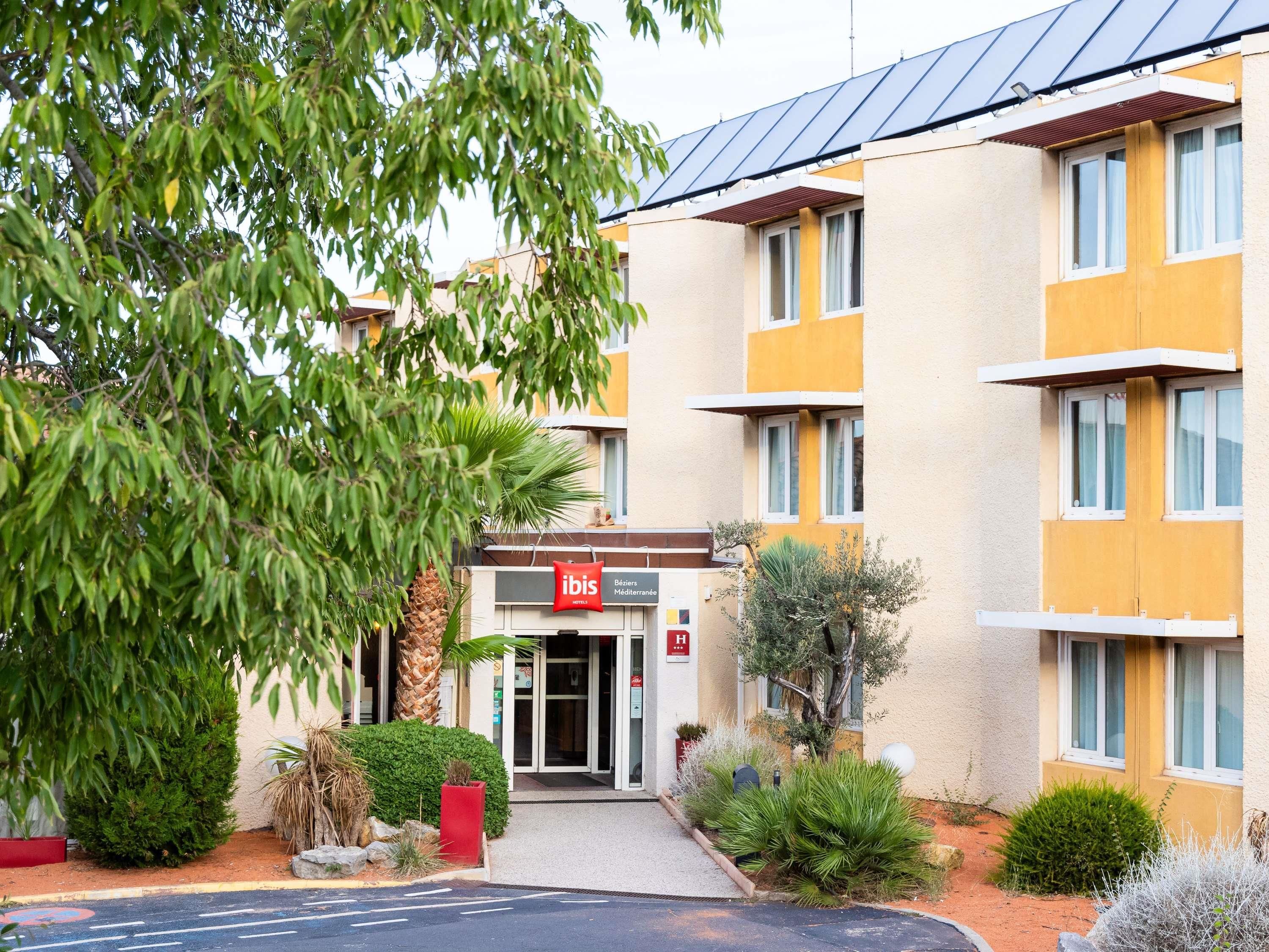 A hotel entrance with a sign for 'ibis', a palm tree, and a small garden area.