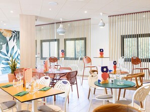 A modern dining area with multiple tables and chairs, a menu stand, and vertical blinds on the windows. - Hôtel ibis Beziers Est Mediterranee (Villeneuve-les-Beziers)