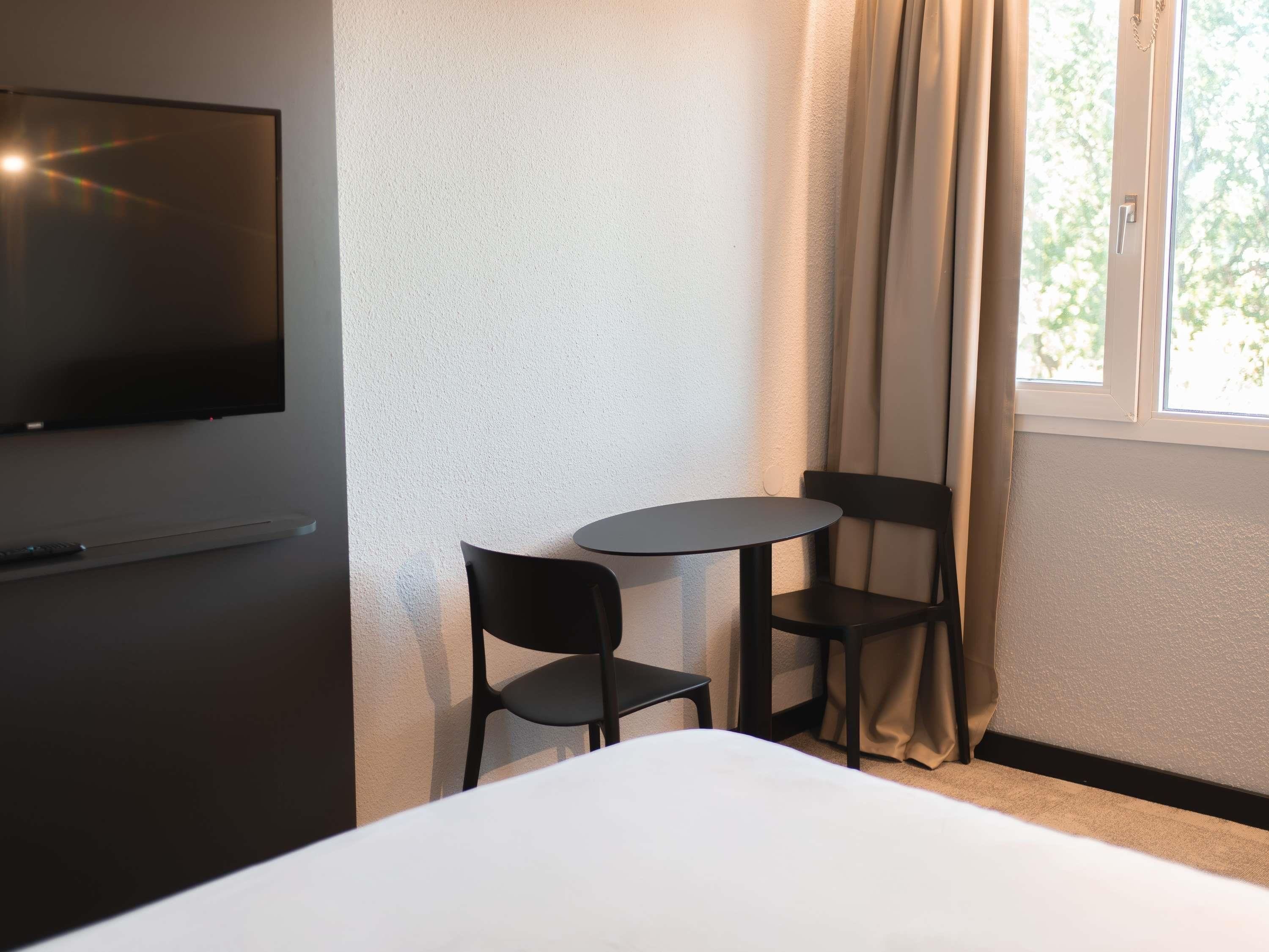 A hotel room with a bed, a TV mounted on the wall, two chairs, a small table, and a window with curtains.