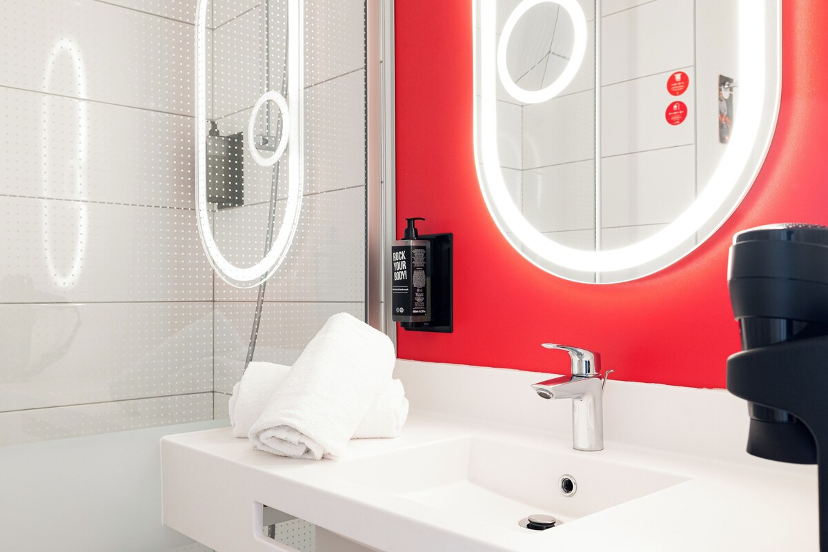 Een moderne badkamer met een witte wastafel, een rode accentmuur en een grote spiegel met een ringlamp.