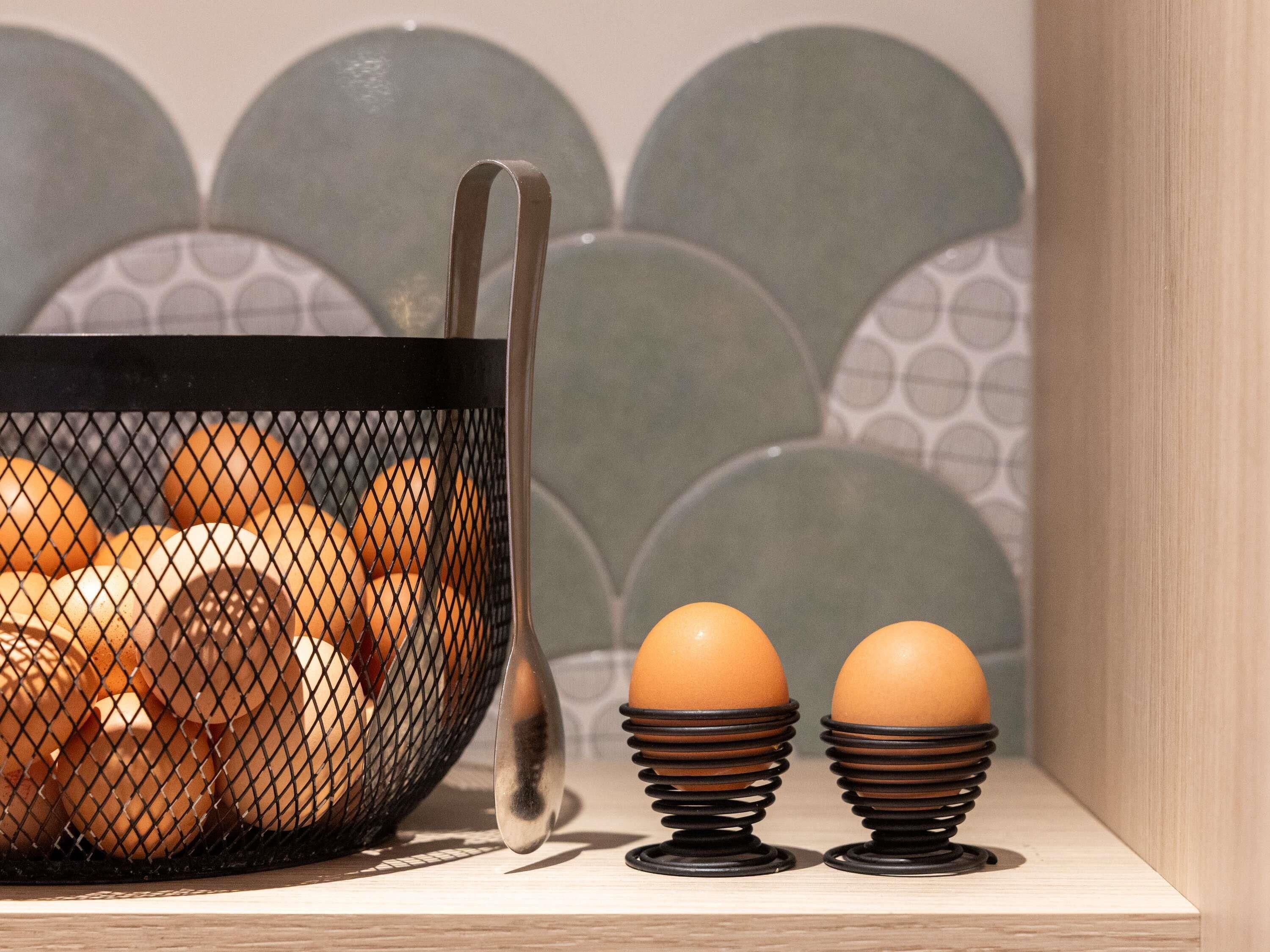 Una encimera de cocina con una cesta de alambre negra que contiene huevos, una cuchara y dos portahuevos.