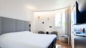 Una habitación de hotel moderna con una cama grande, una mesita con dos sillas, un televisor y una ventana con cortinas.