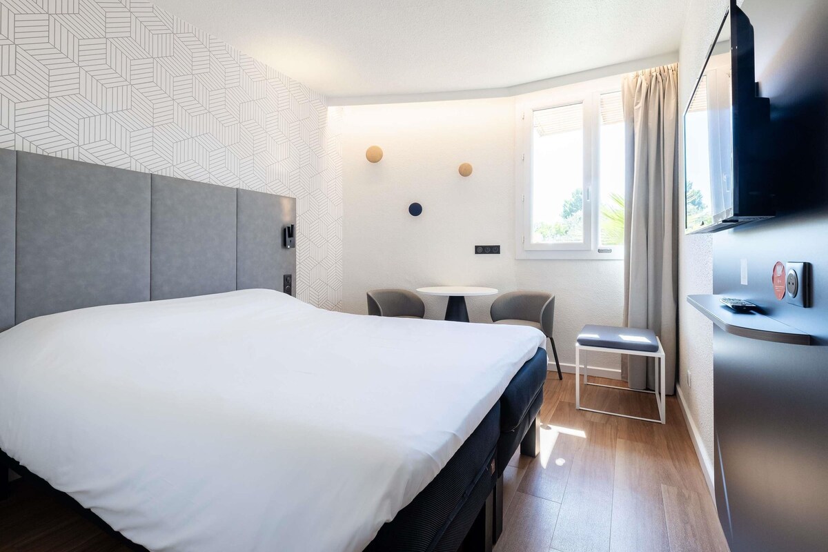 Een moderne hotelkamer met een groot bed, een klein tafeltje met twee stoelen, een televisie en een raam met gordijnen.