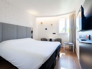 Een moderne hotelkamer met een groot bed, een klein tafeltje met twee stoelen, een televisie en een raam met gordijnen.