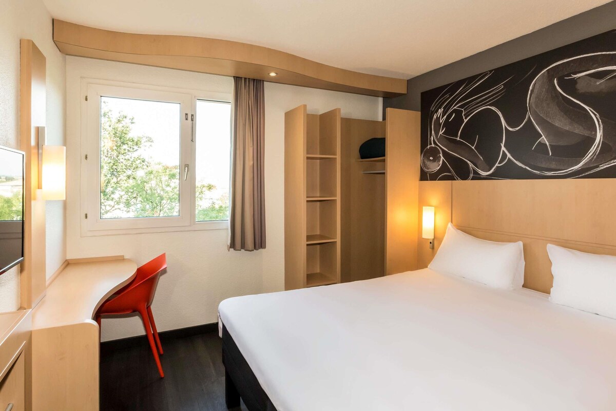 Een hotelkamer met een bed, een bureau, een stoel, een kledingkast en een raam met gordijnen.