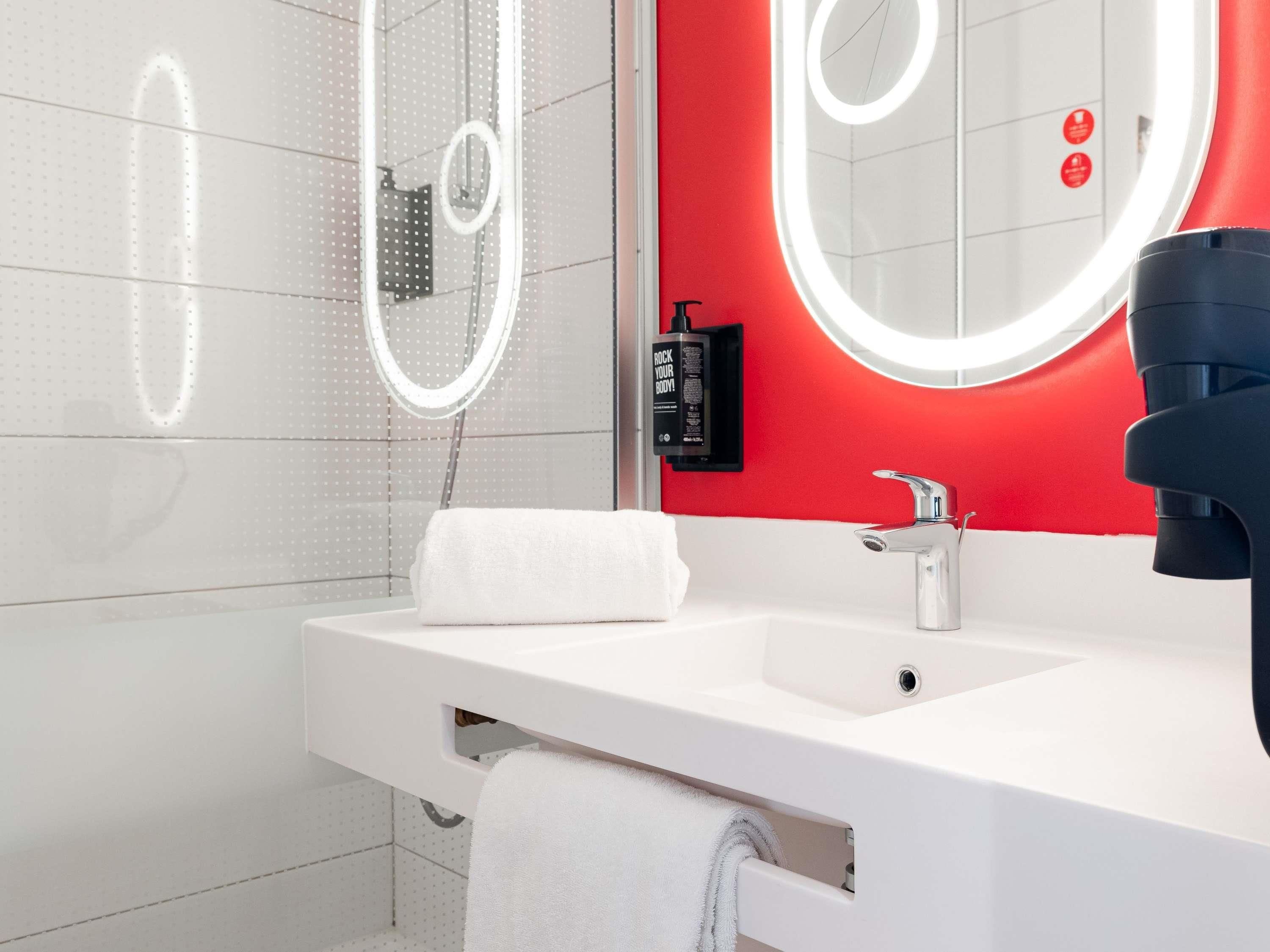 A modern bathroom with a white sink, a mirror with a circular light, and a red accent wall.