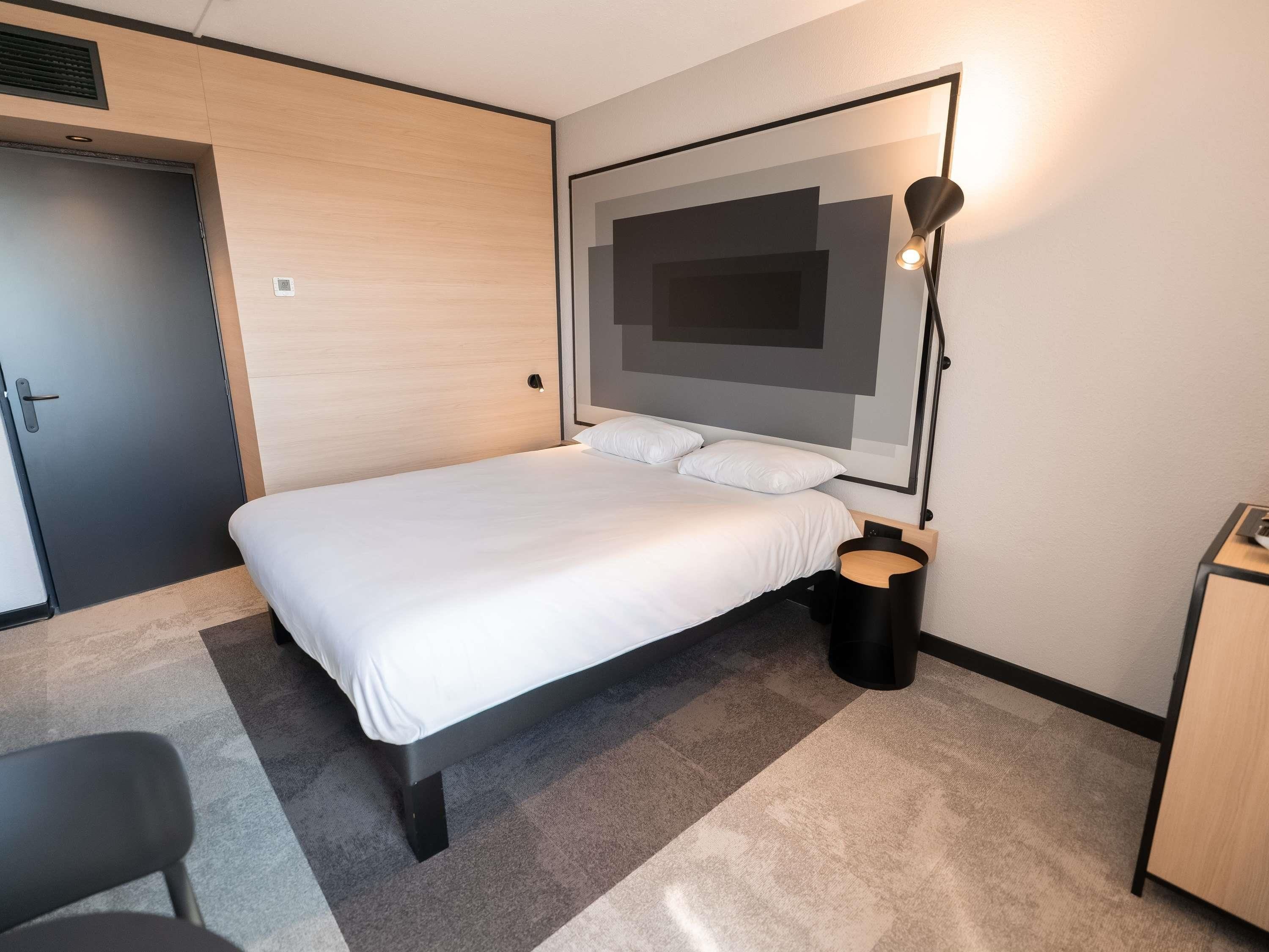 Una habitación de hotel moderna con una cama, un escritorio, una silla, un armario y una lámpara fijada a la pared.