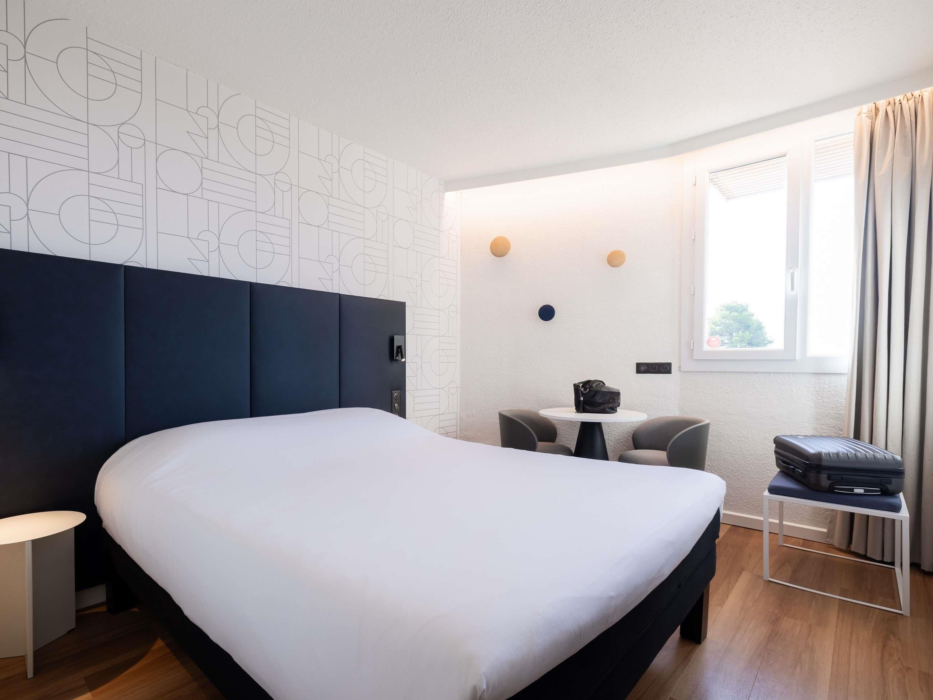 Een moderne hotelkamer met een groot bed, een klein tafeltje en een stoel. Er is een raam met gordijnen en uitzicht op buiten.