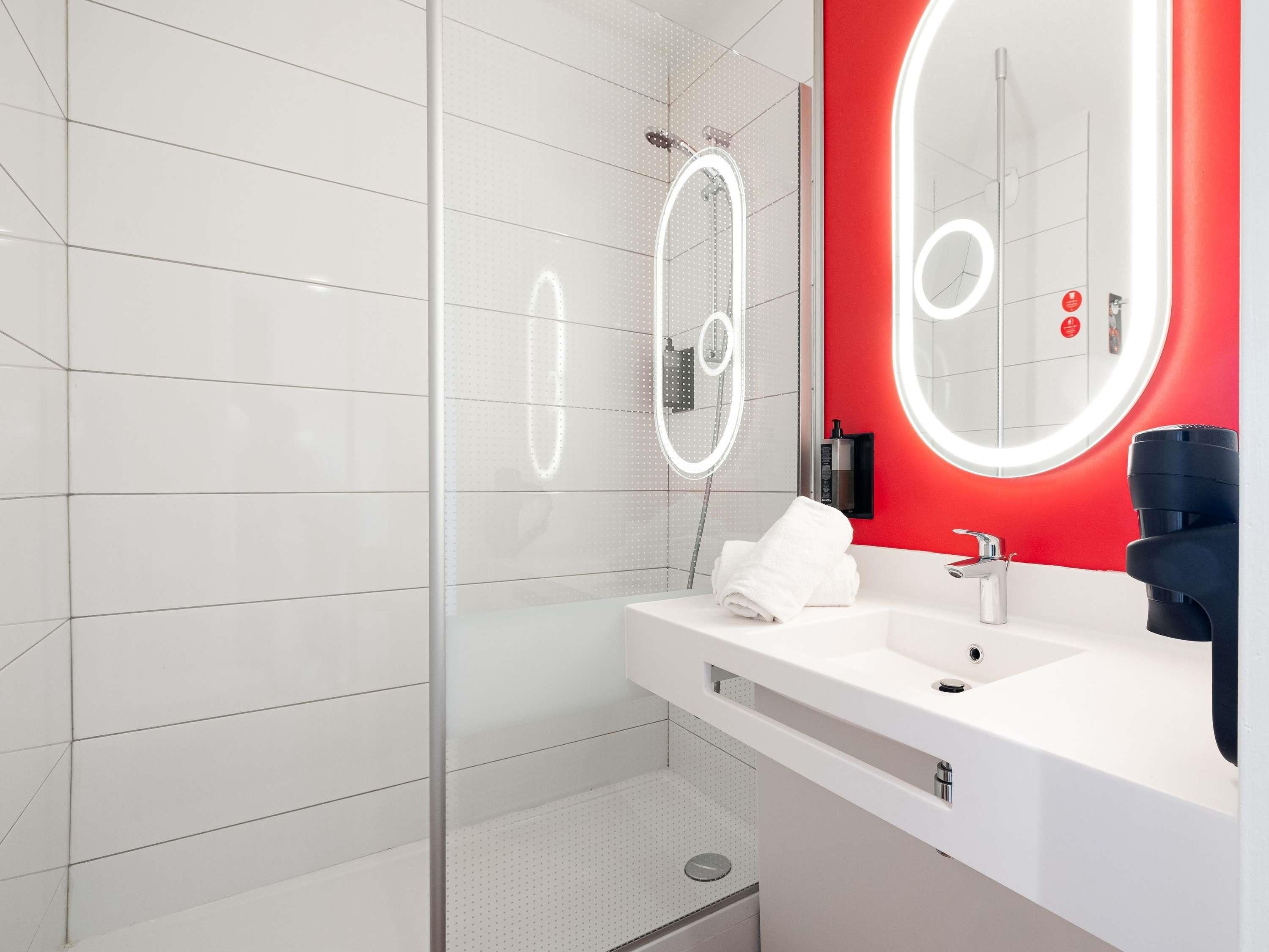 A modern bathroom with a white tiled shower area, a white sink with a red accent wall, and a large mirror with illuminated edges.