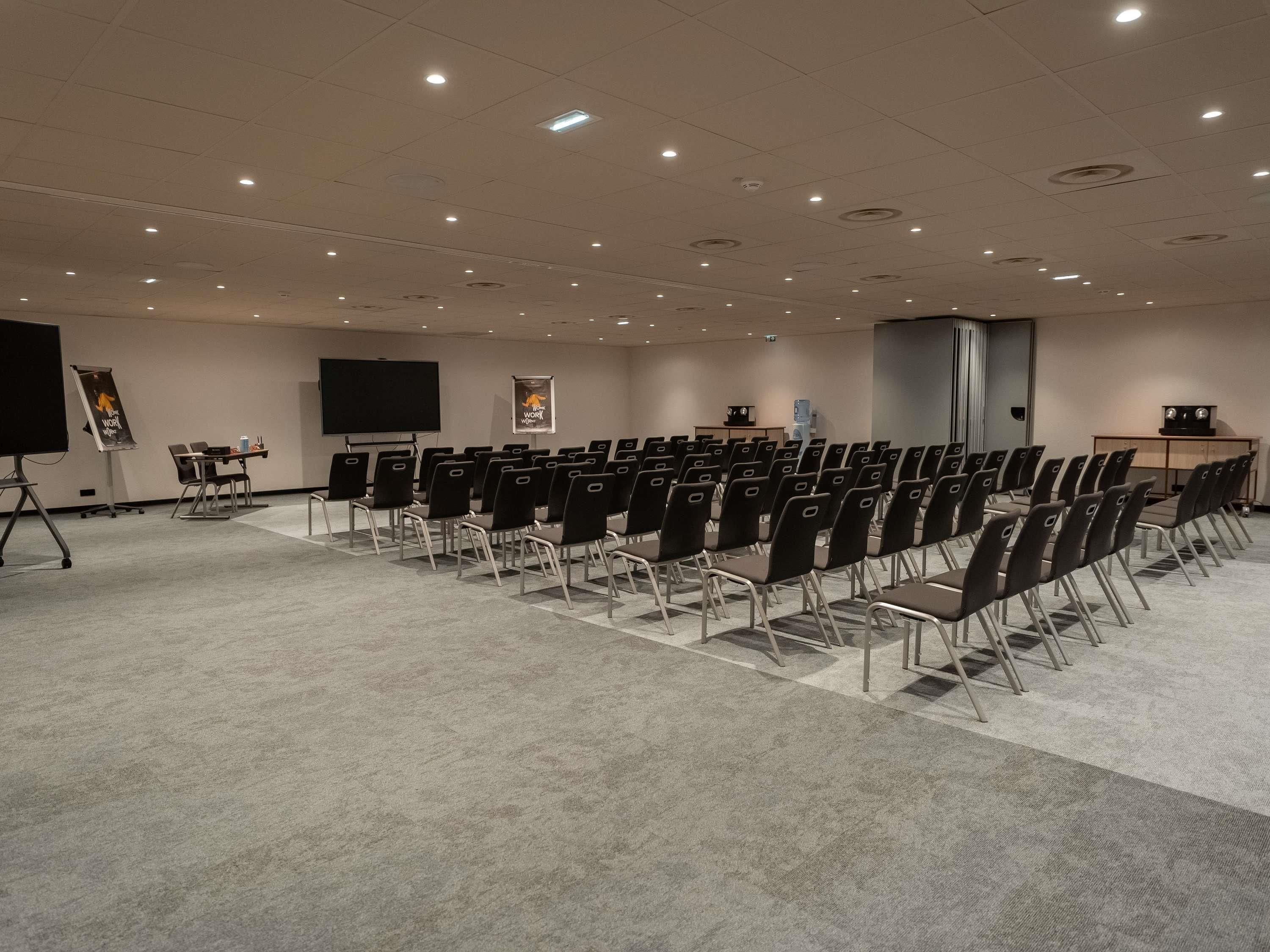 A conference room with rows of chairs, a projector screen, a presentation screen, and a water dispenser.