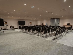 Un salón de conferencias con filas de sillas, una pantalla de proyector, una pantalla para presentaciones y un dispensador de agua.