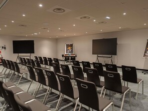 Une salle de conférence avec des rangées de chaises, deux grands écrans, un pupitre et un projecteur.