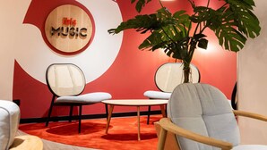 A room with a red wall, a circular wooden sign with 'iBis MUSIC' text, two wicker chairs, a round table, and a potted plant. - Hôtel ibis Beziers Est Mediterranee (Villeneuve-les-Beziers)