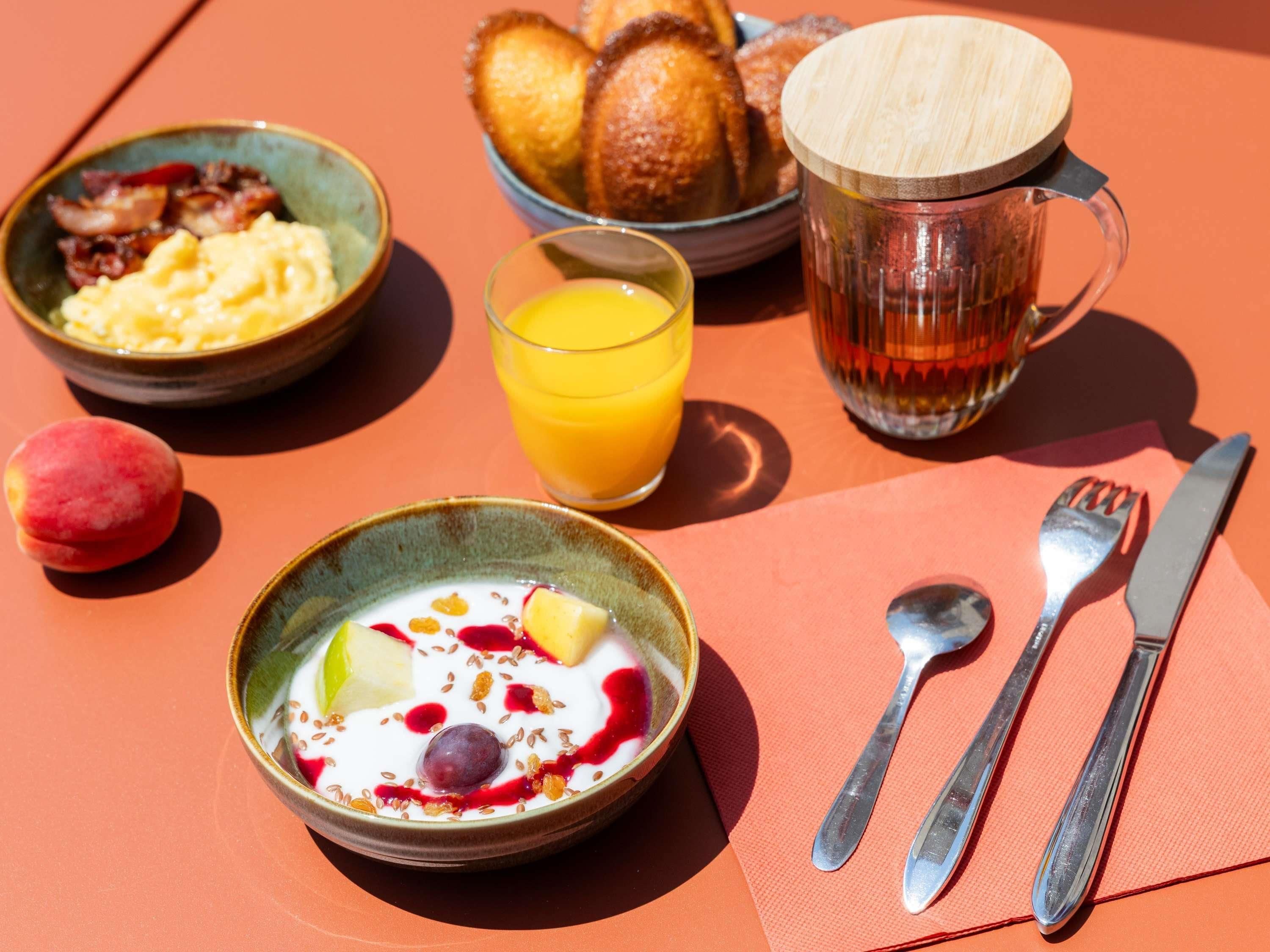 A breakfast setting with a bowl of yogurt topped with fruits and seeds, a glass of orange juice, a mug of tea, and a plate of croissants.
