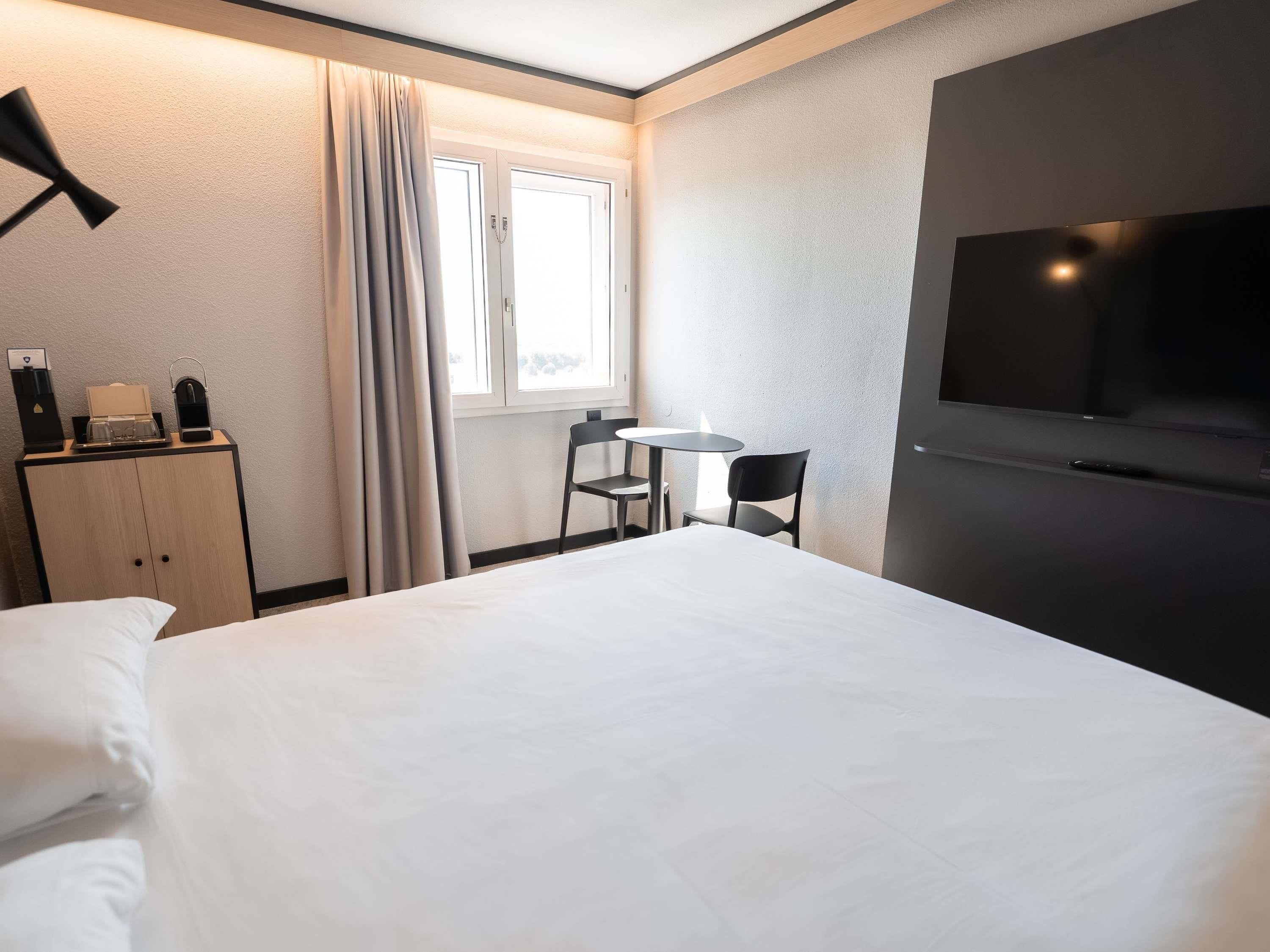 A hotel room with a bed, a desk with a chair, a television, and a window with curtains.