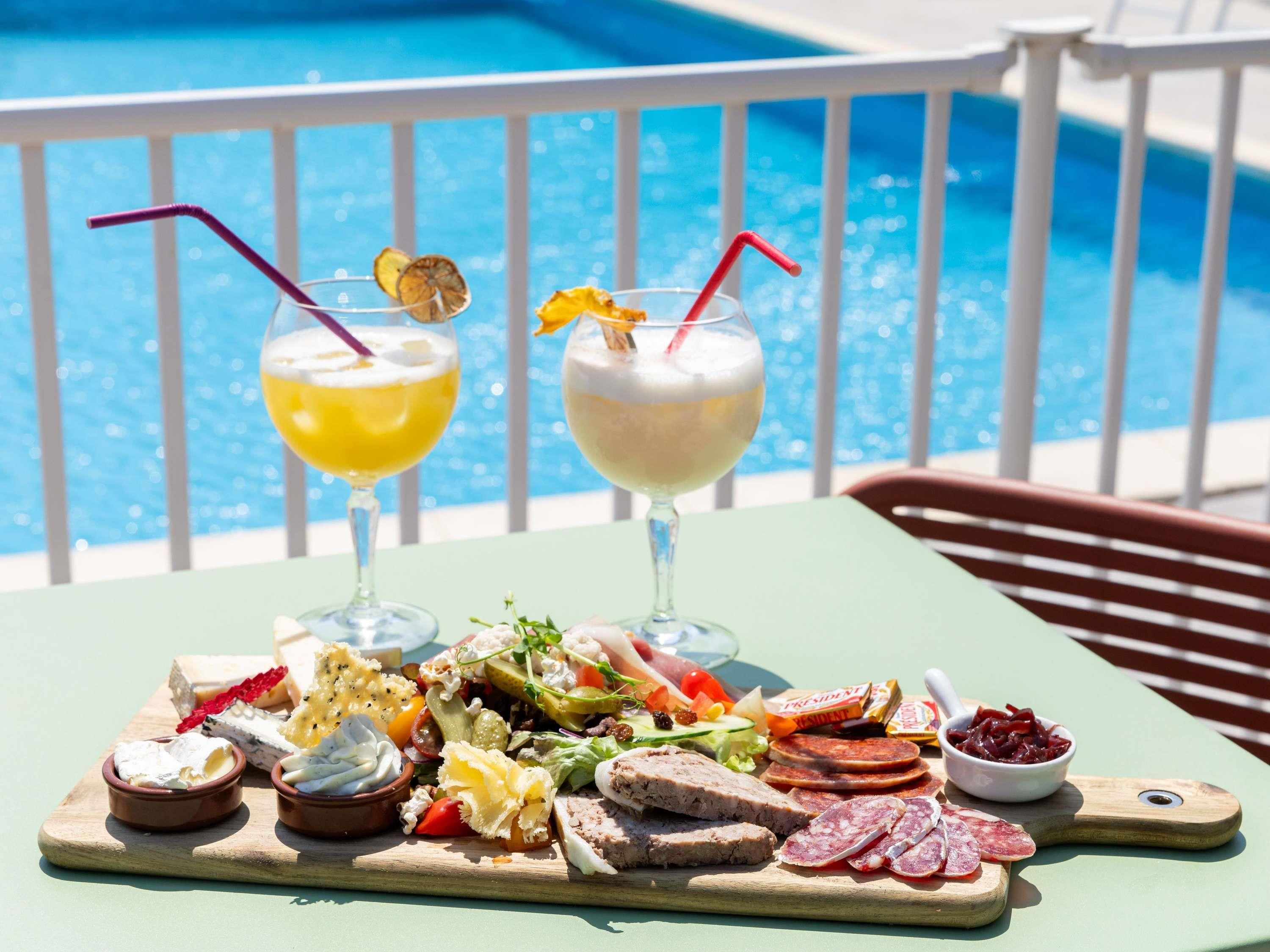 Ein Tisch am Pool mit einer Wurstplatte, zwei Getränken mit Trinkhalmen und einem Pool im Hintergrund.