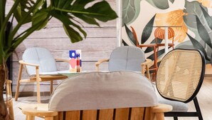 Un coin salon accueillant avec des chaises en bois et un fauteuil en rotin, une grande plante à feuilles luxuriantes et un tableau abstrait aux couleurs vives accroché au mur.