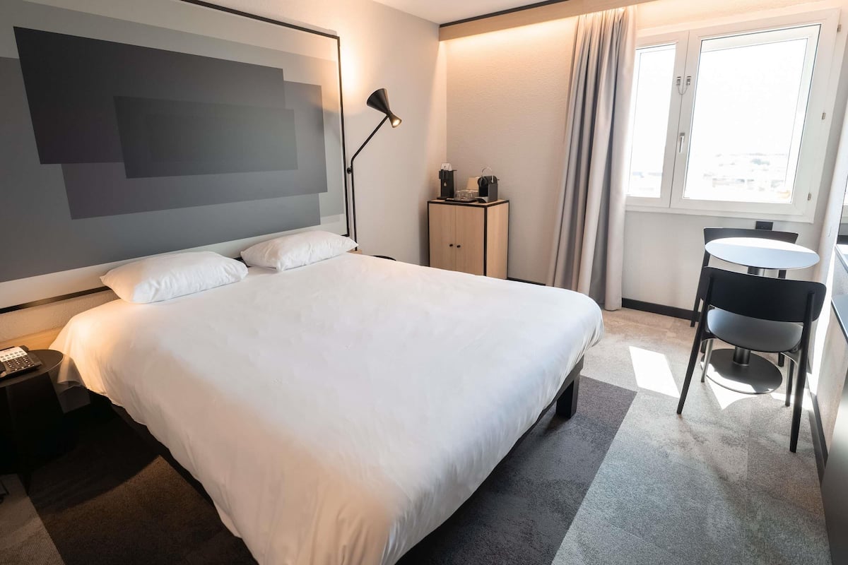 Une chambre d’hôtel avec un grand lit, un bureau avec une chaise, une lampe, une armoire et une fenêtre avec des rideaux.