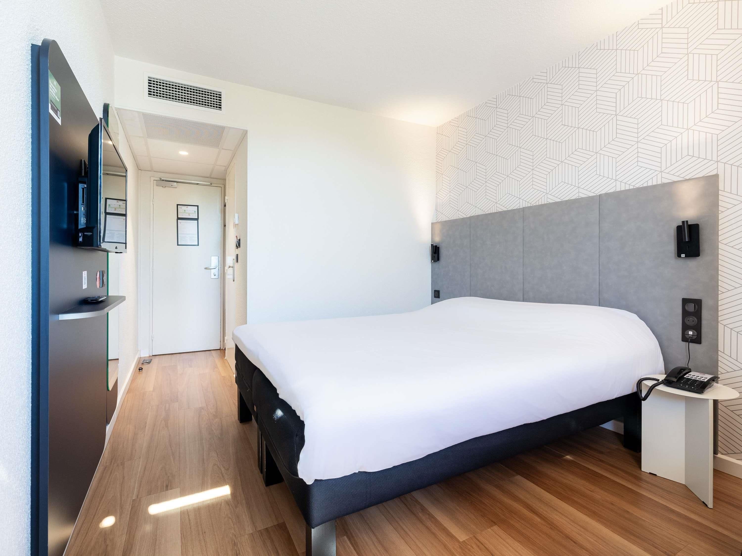 A modern hotel room with a large bed, a flat-screen TV, a small bedside table, and a patterned wall.
