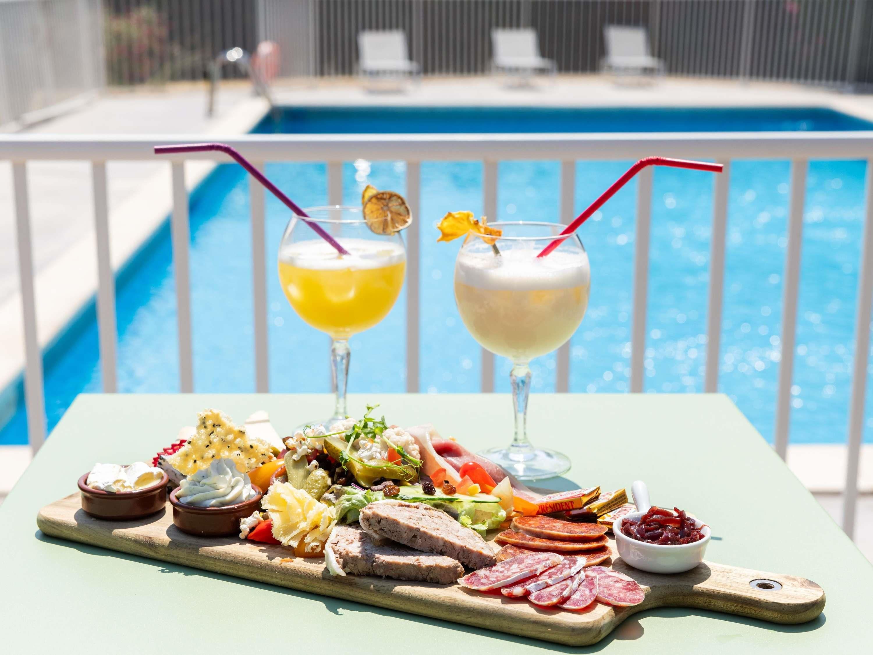 Un tavolo a bordo piscina con un tagliere di salumi, due bevande e vista sulla piscina.