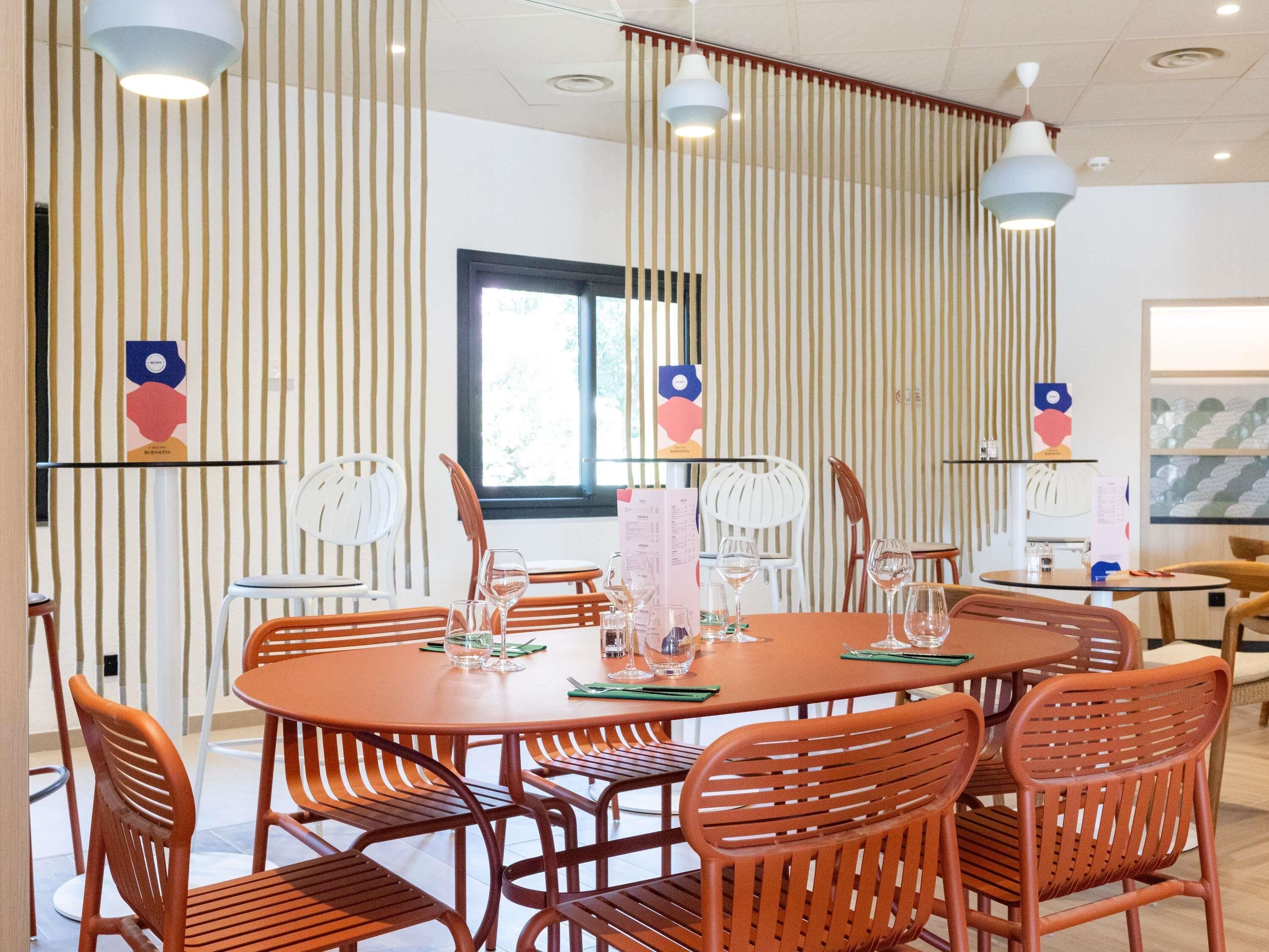 A modern dining area with round tables and orange chairs, featuring vertical striped walls and pendant lighting.