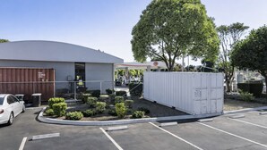 Un estacionamiento con un edificio blanco, un árbol y un área cercada con un contenedor blanco.