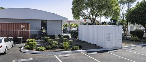 A parking lot with a white building, a tree, and a fenced area with a white container.