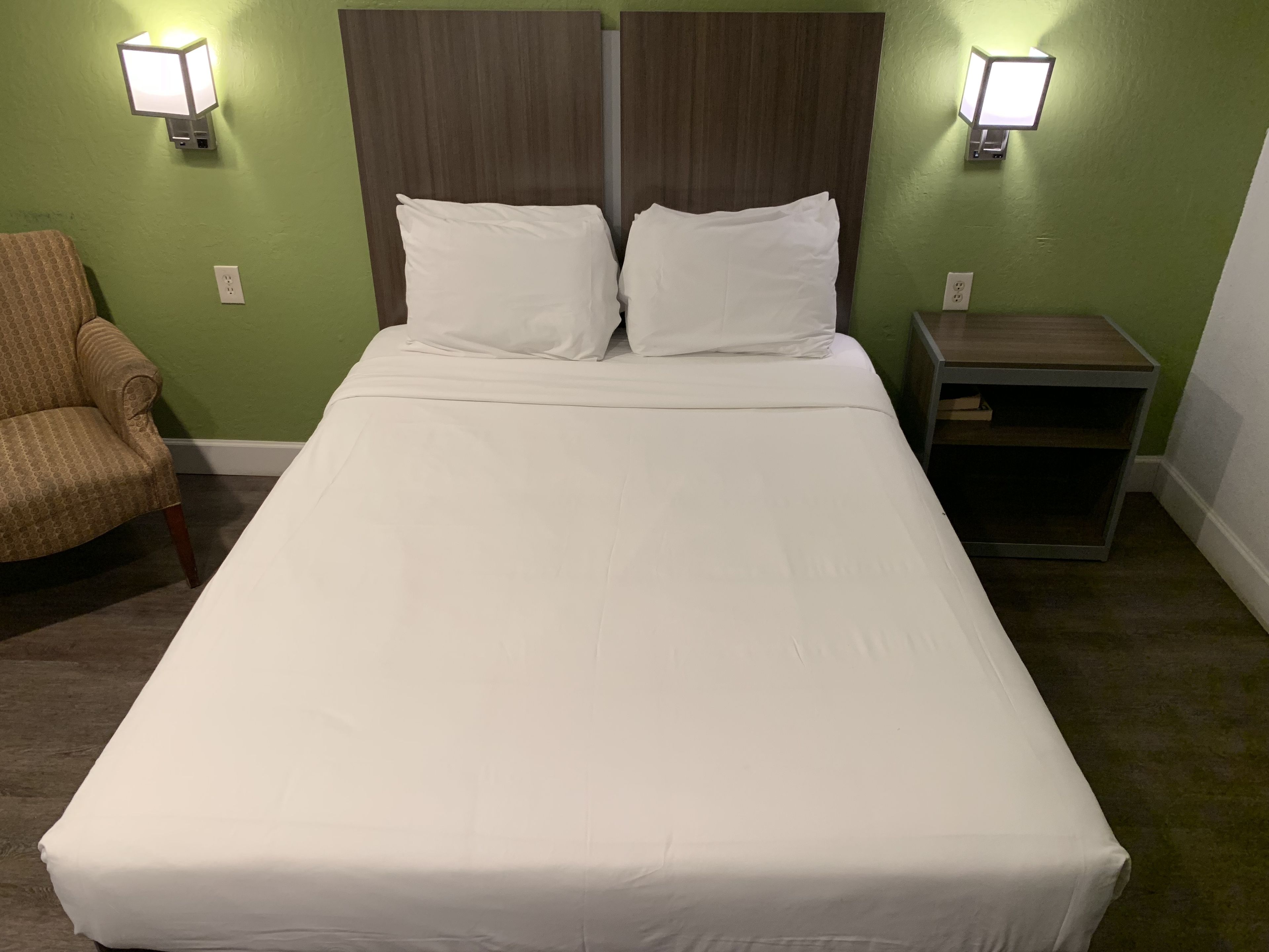 Una cama bien tendida con sábanas blancas en una habitación con paredes verdes y dos mesitas de noche fijadas a la pared.
