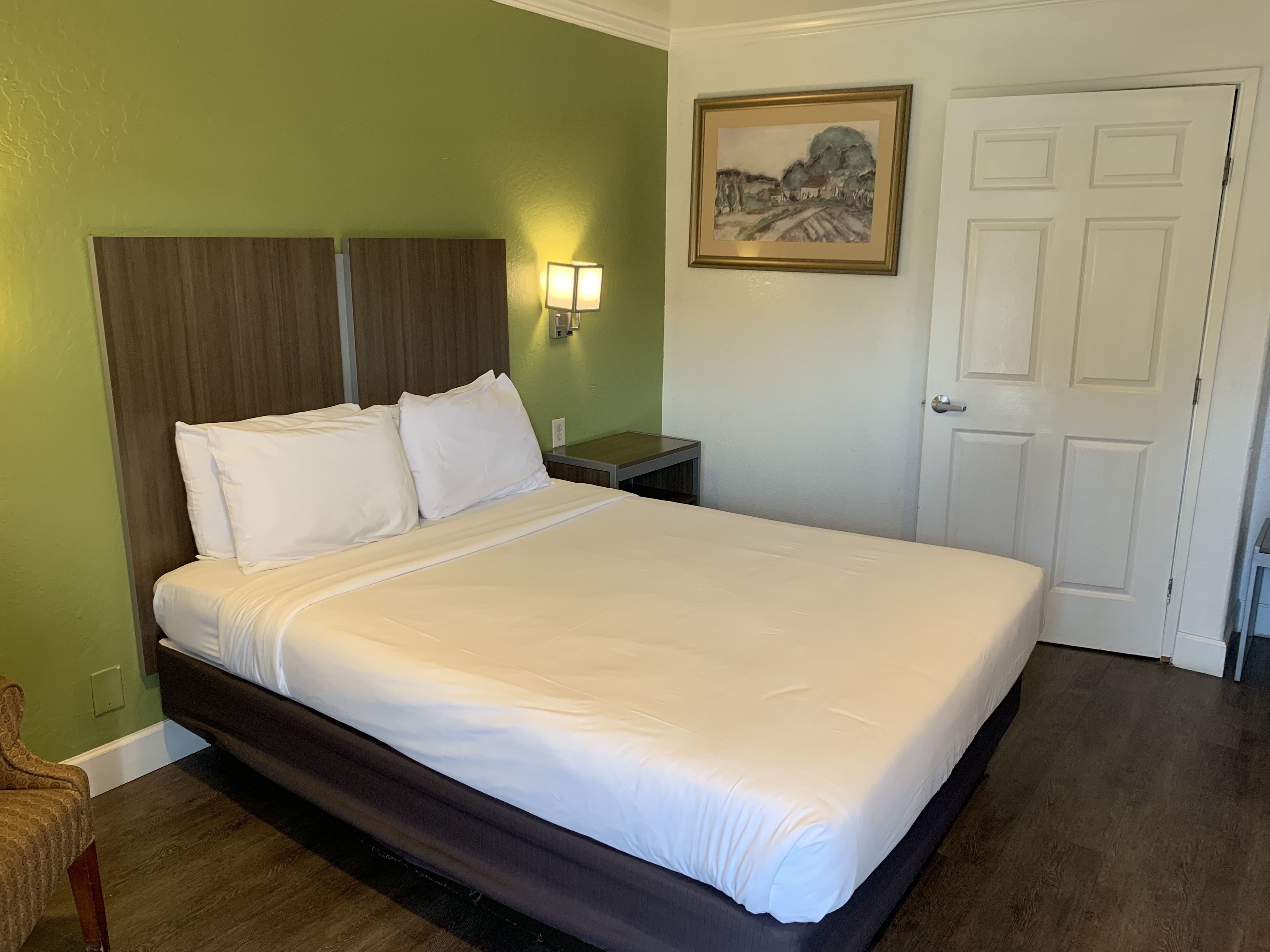 A neatly made bed with white linens in a room with green walls, a wooden headboard, a nightstand with a lamp, and a door with a framed picture above it.