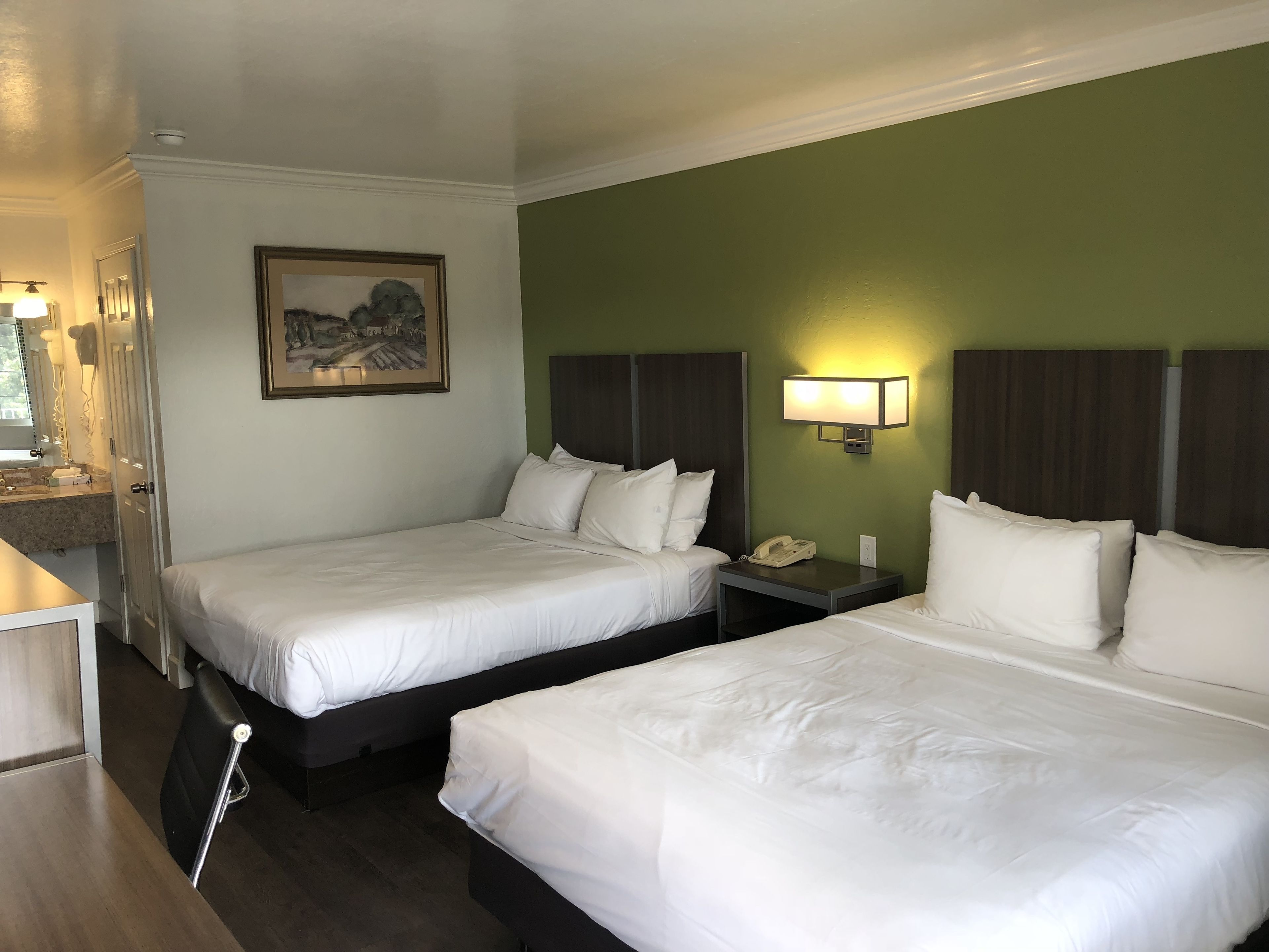 Habitación de hotel con dos camas, una mesita de noche con teléfono, un cuadro en la pared y un baño con espejo e iluminación.