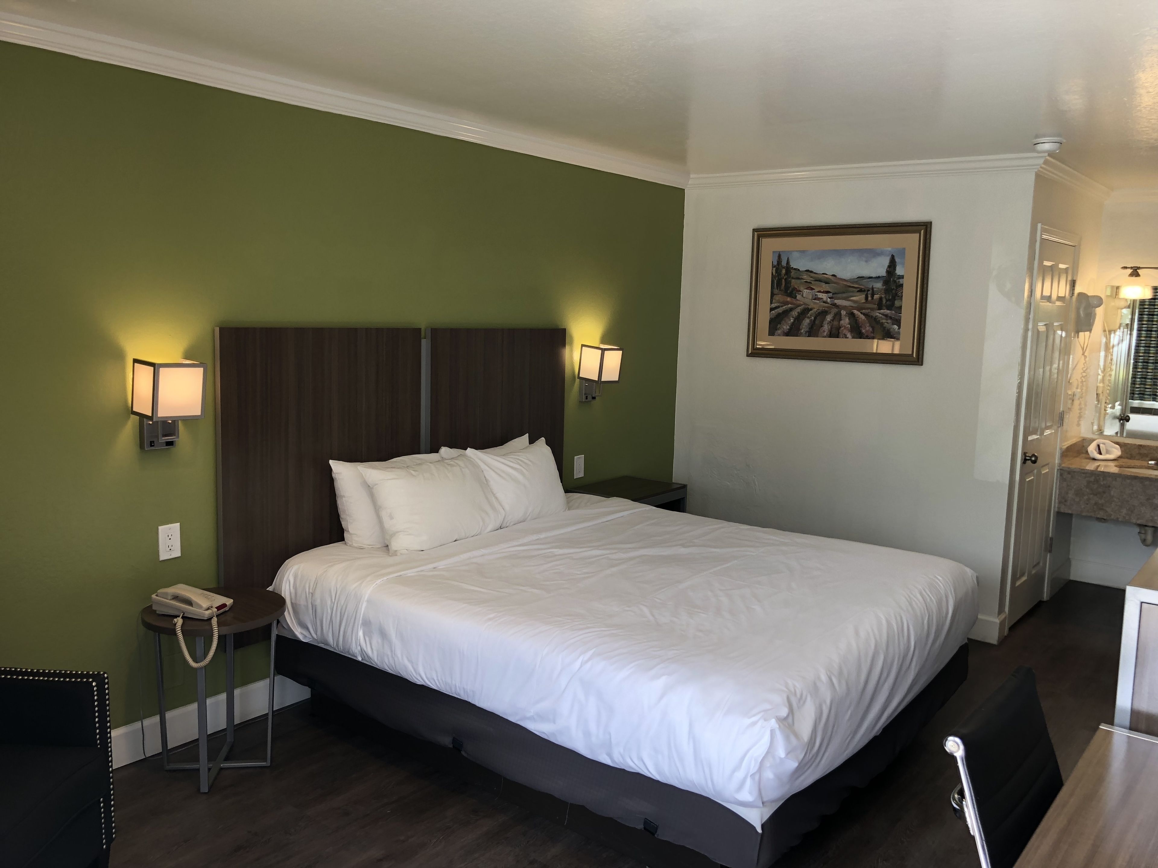 A hotel room with a large bed, two bedside lamps, a painting on the wall, a telephone on a side table, and a mirror above the sink.