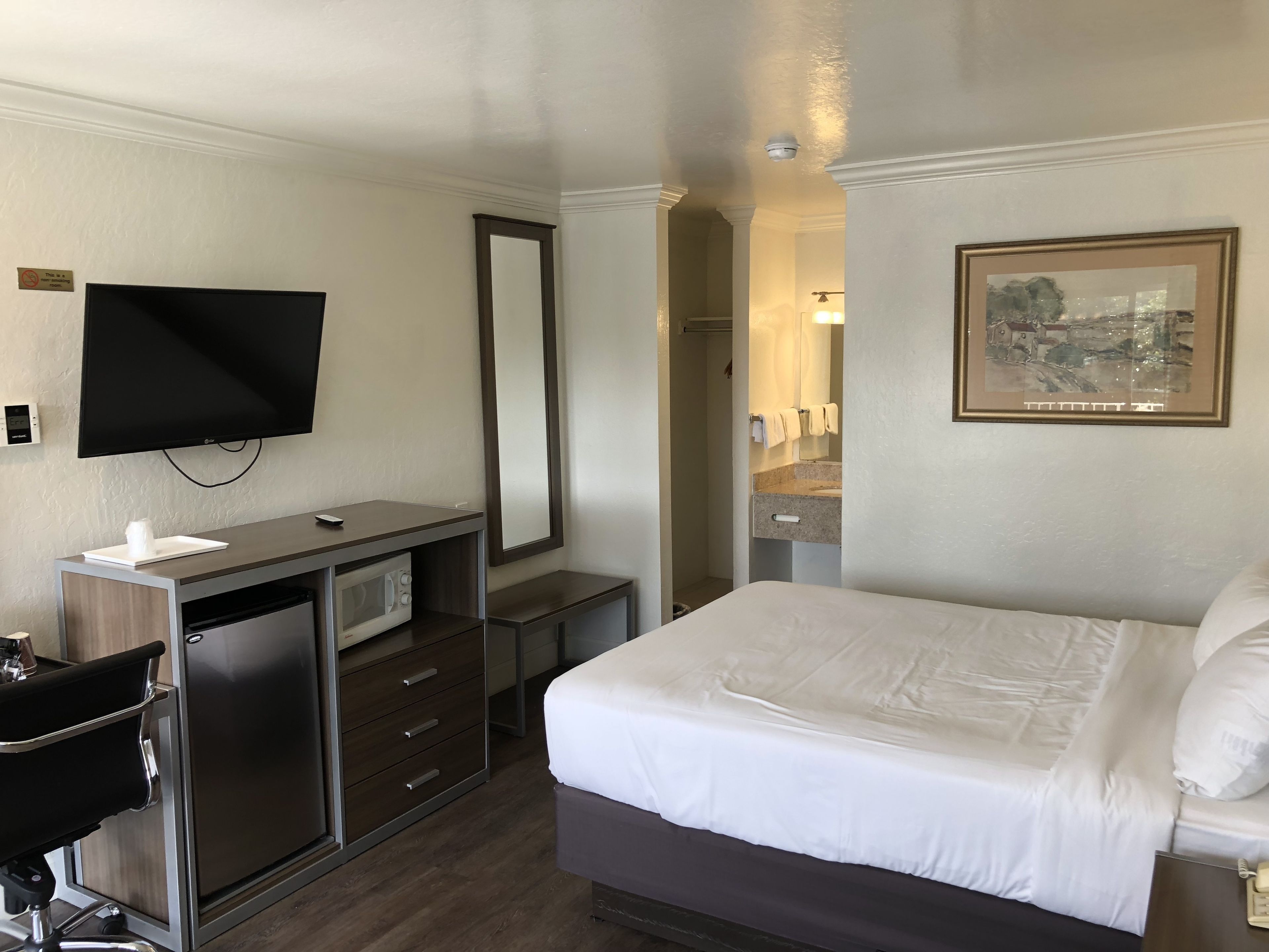 A hotel room with a bed, a desk with a computer, a television, a mirror, a bathroom, and a framed picture on the wall.