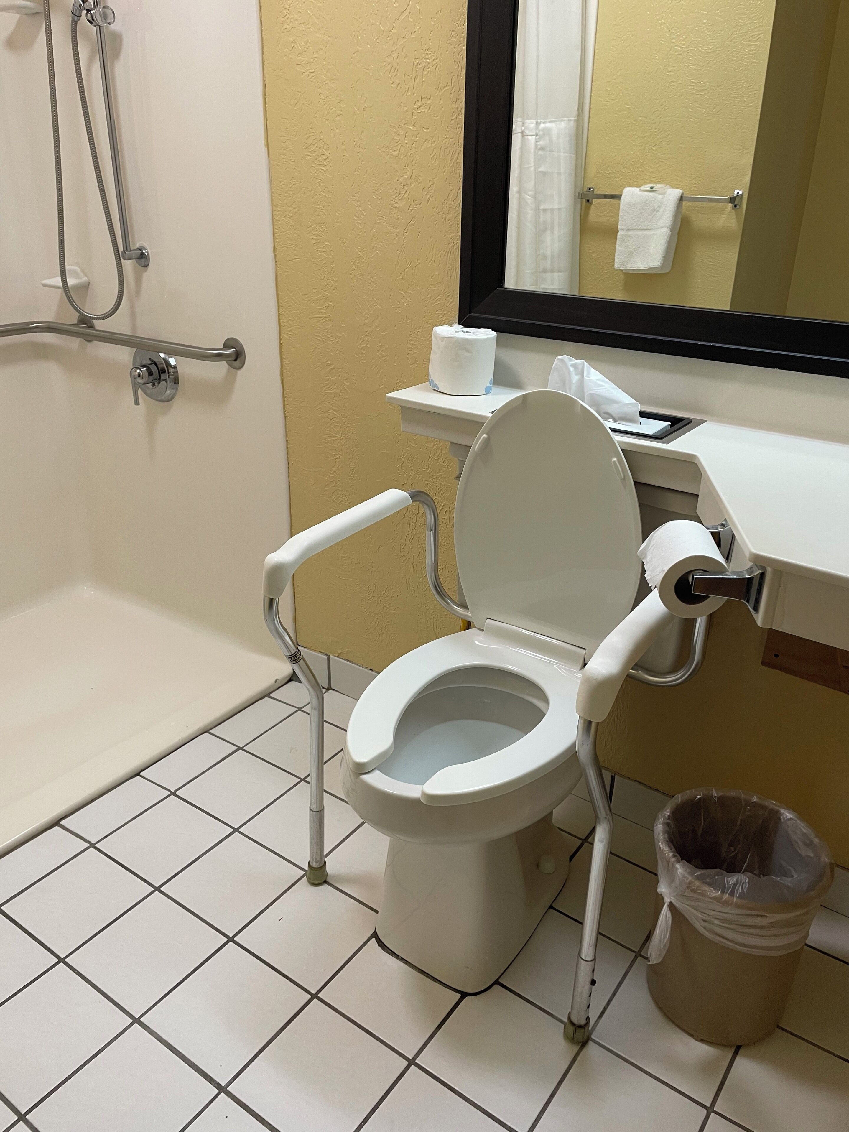 A bathroom with a white toilet, a grab bar, a shower, and a trash can.