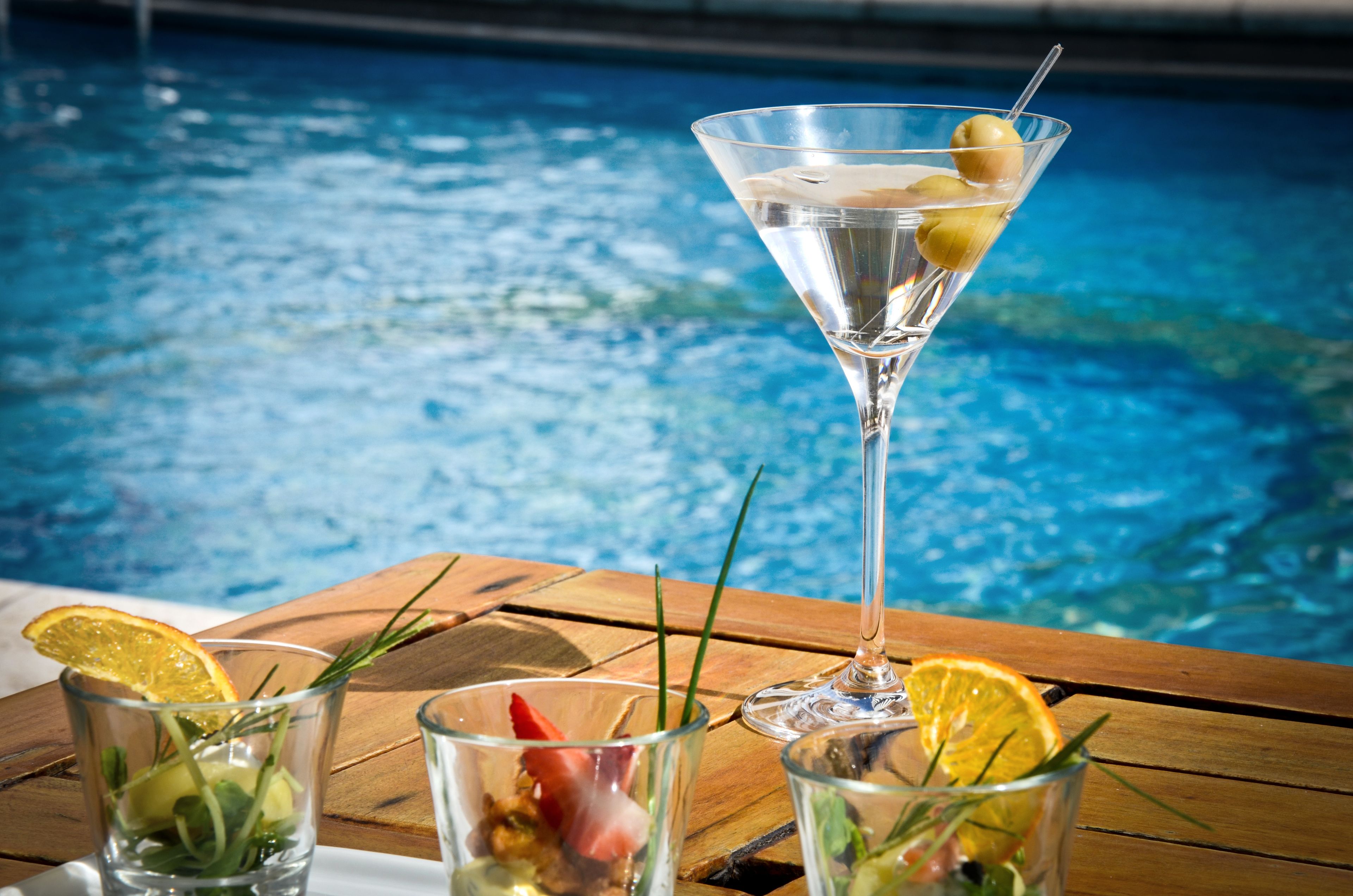 Una mesa junto a la piscina con tres vasos de bebidas, uno de martini, y una piscina al fondo.