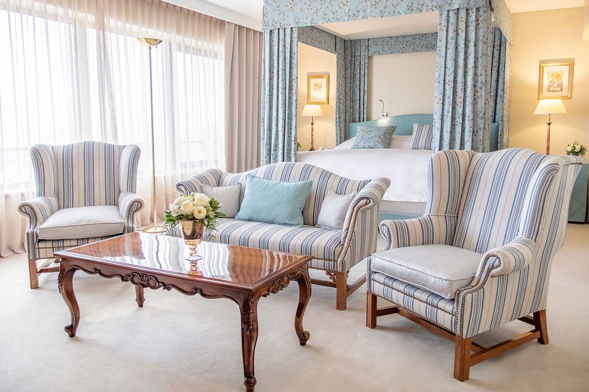 A hotel room with a large bed, two striped armchairs, a wooden coffee table, and a floral arrangement.