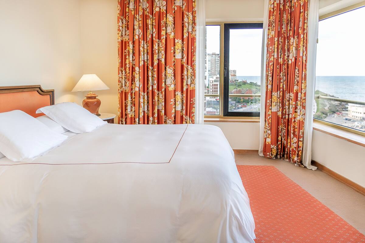 A hotel room with a bed, a nightstand, a lamp, floral curtains, and a view of the sea.