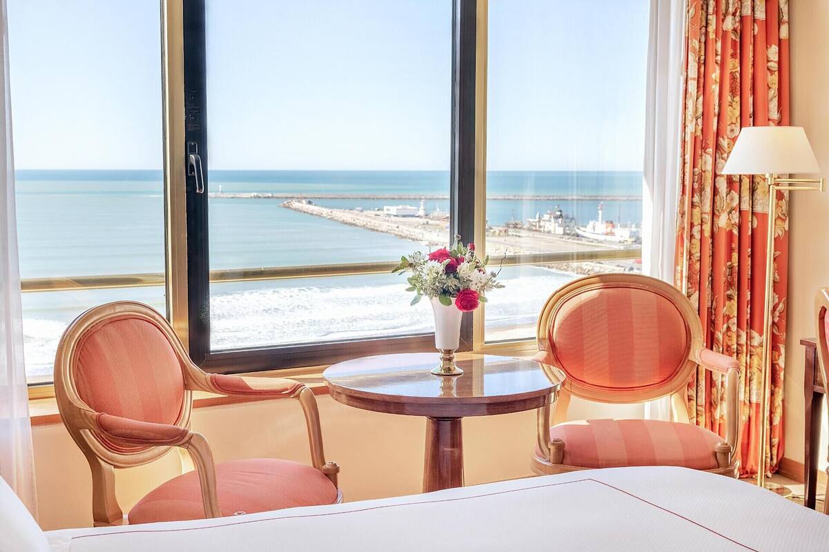 A hotel room with a large window overlooking a beach, a round table with chairs, and a vase with flowers.