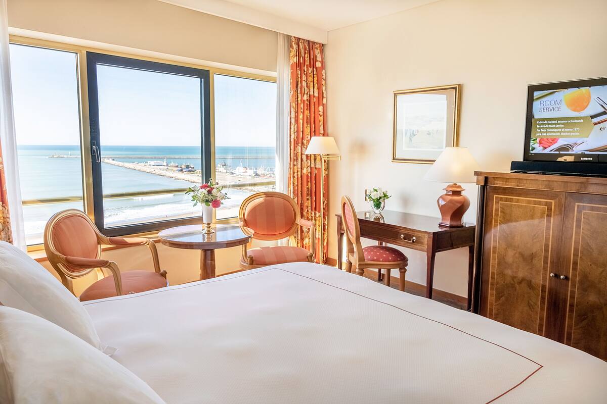 A hotel room with a bed, two chairs, a small table with a vase, a TV on a cabinet, and a view of the sea.