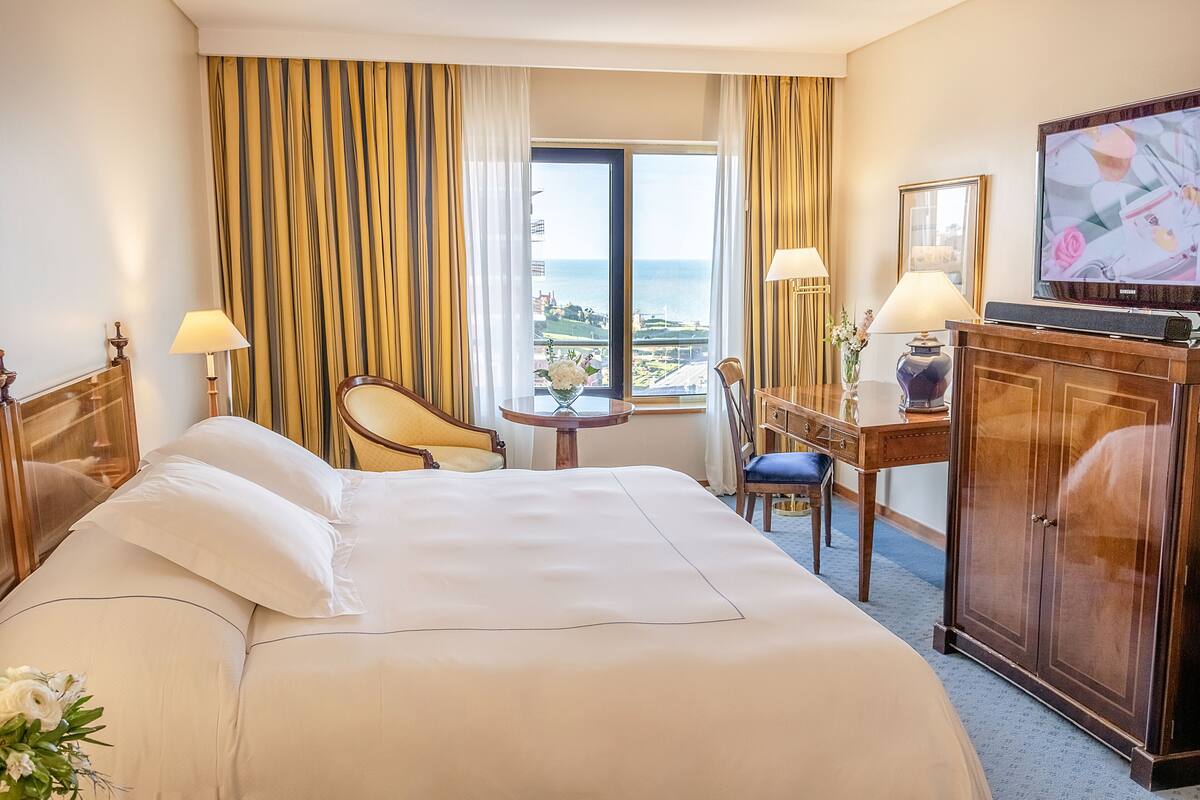 A hotel room with a large bed, a desk, a chair, a television, and a view of the sea.