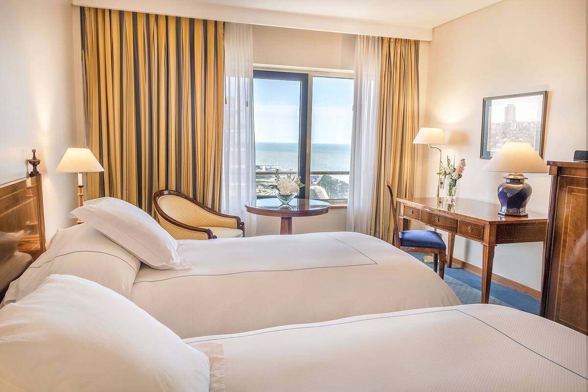 A hotel room with two beds, a desk, a chair, a table, and a view of the sea.