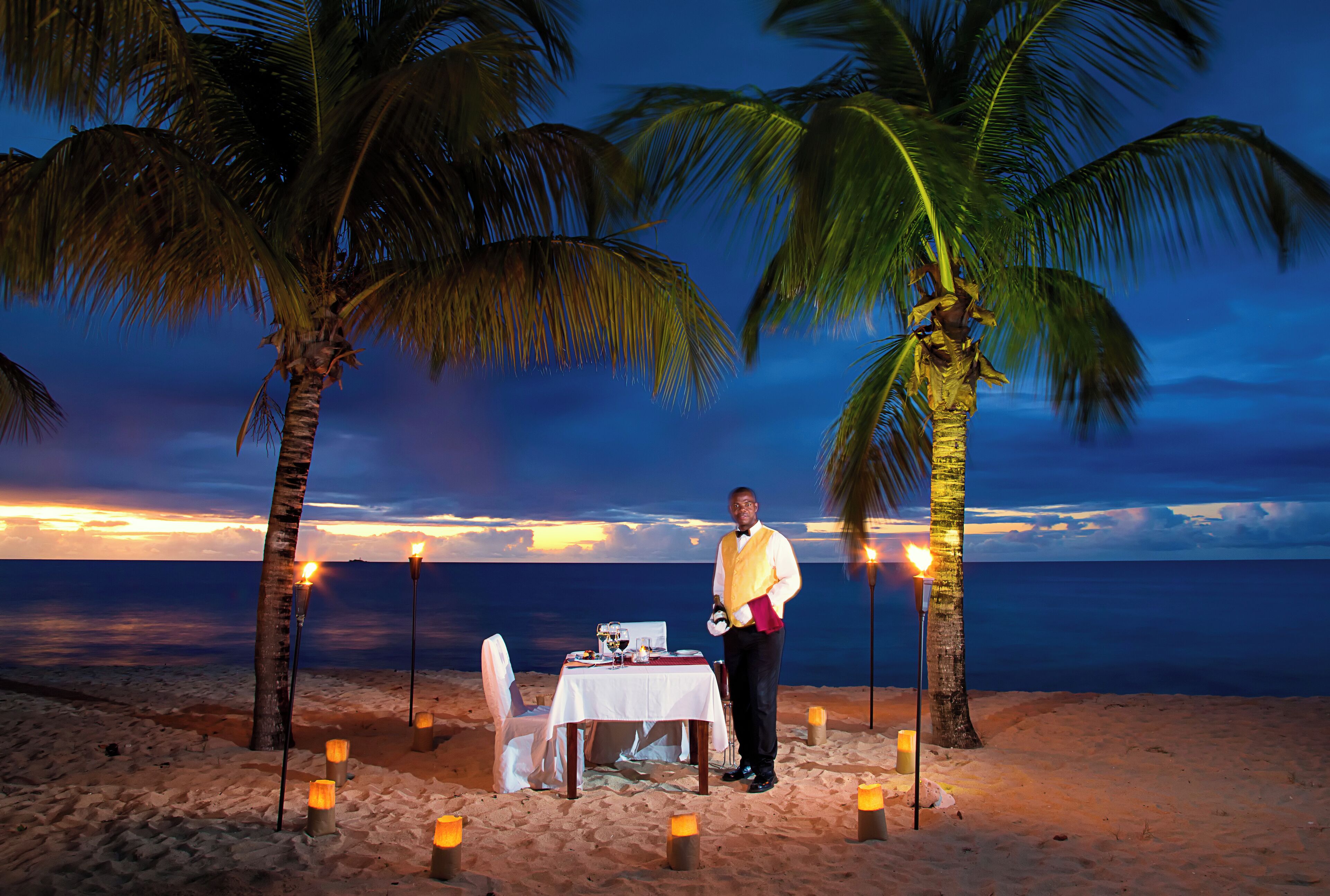 Cena a la orilla de la playa con una mesa, sillas y velas, rodeada de palmeras y vista al océano al atardecer.