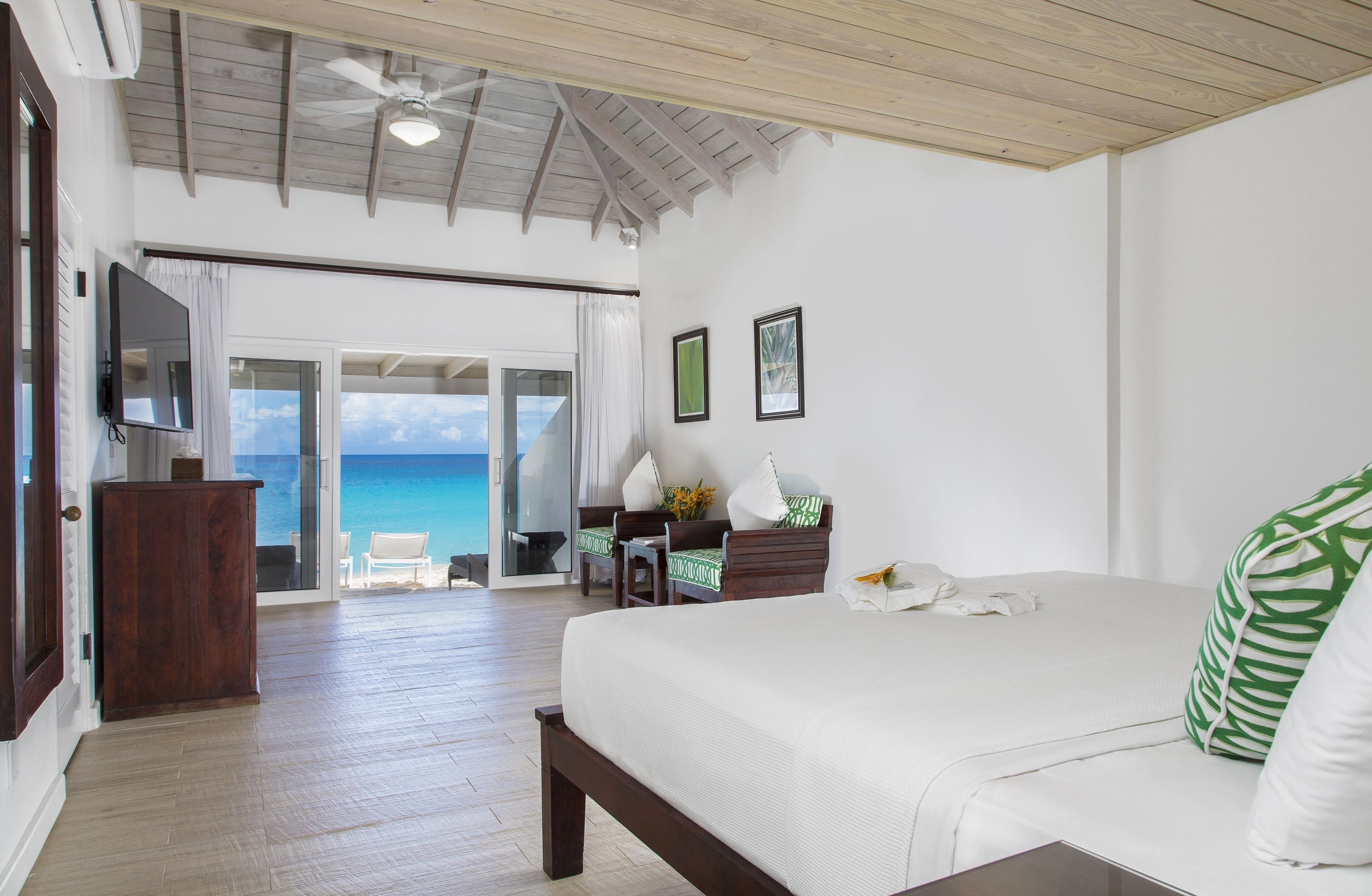 A spacious bedroom with a large bed, a wooden ceiling, a television, and a view of the ocean through a sliding door.