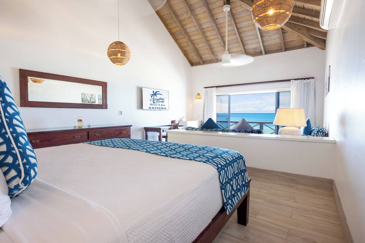 A bedroom with a large bed, a window with a view of the ocean, a ceiling fan, and a wall-mounted mirror.