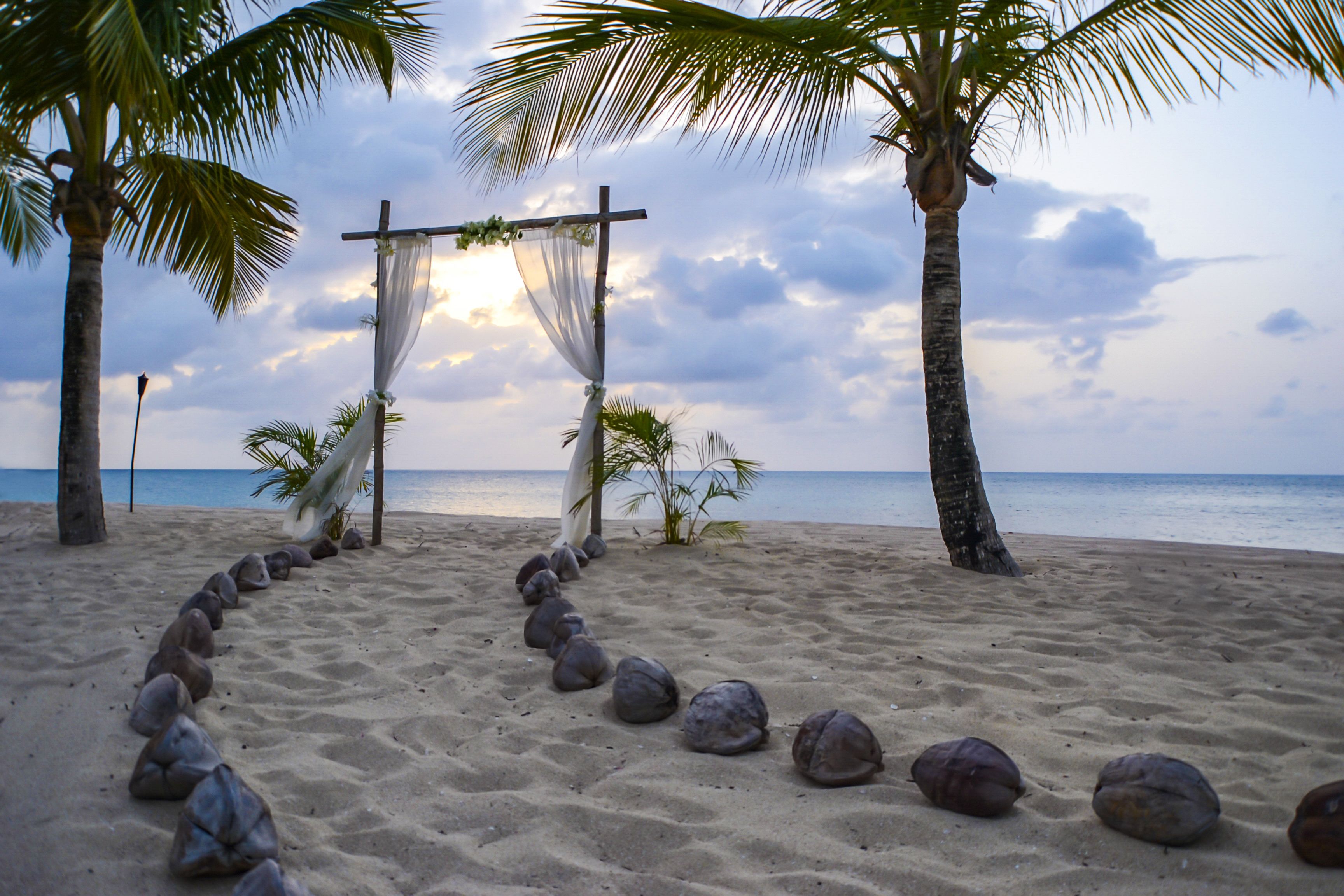 A beach with a pathway lined with rocks leading to a wooden archway adorned with white fabric and greenery.
